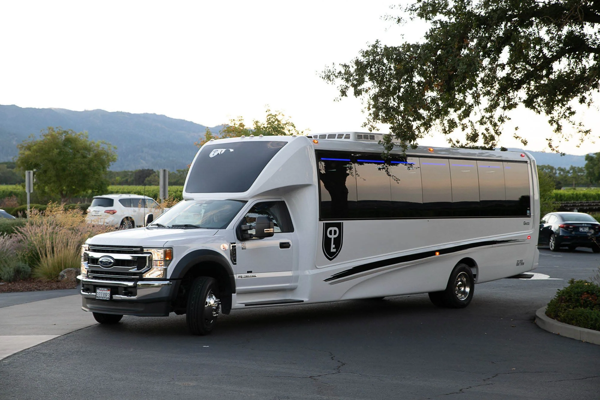 A white autonomous delivery vehicle driving on a street in a suburban or park area during daytime, with trees and mountains in the background.