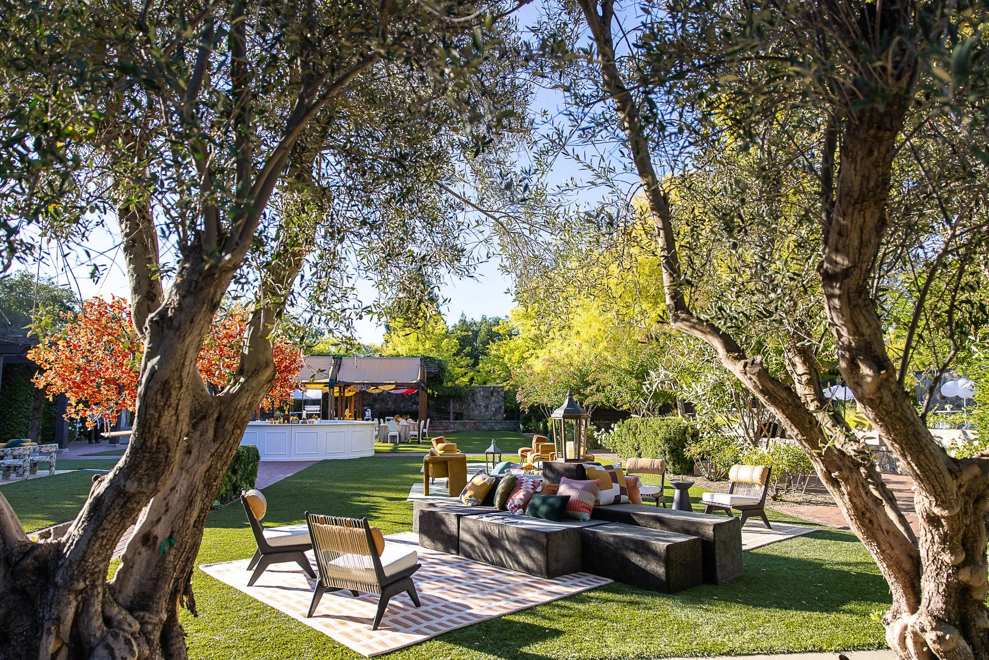 An outdoor garden party setup with lounge chairs, tables, and an overhang. Tall trees with green leaves frame the view, and colorful trees add fall foliage. The scene features a well-maintained lawn, decorative lanterns, and a sunny sky.