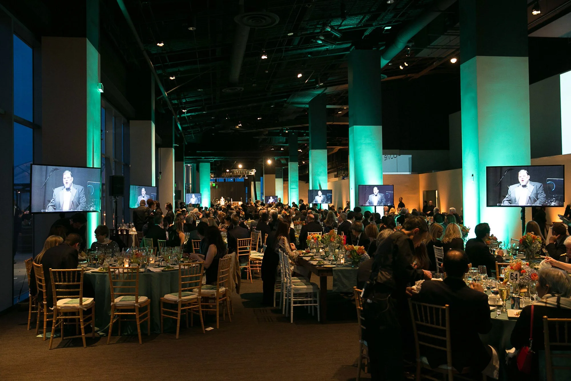 A large conference or banquet hall filled with many round tables decorated with floral centerpieces and set with glassware and gold chairs. There are numerous attendees seated. Multiple screens display a man in a suit speaking at a podium on stage in