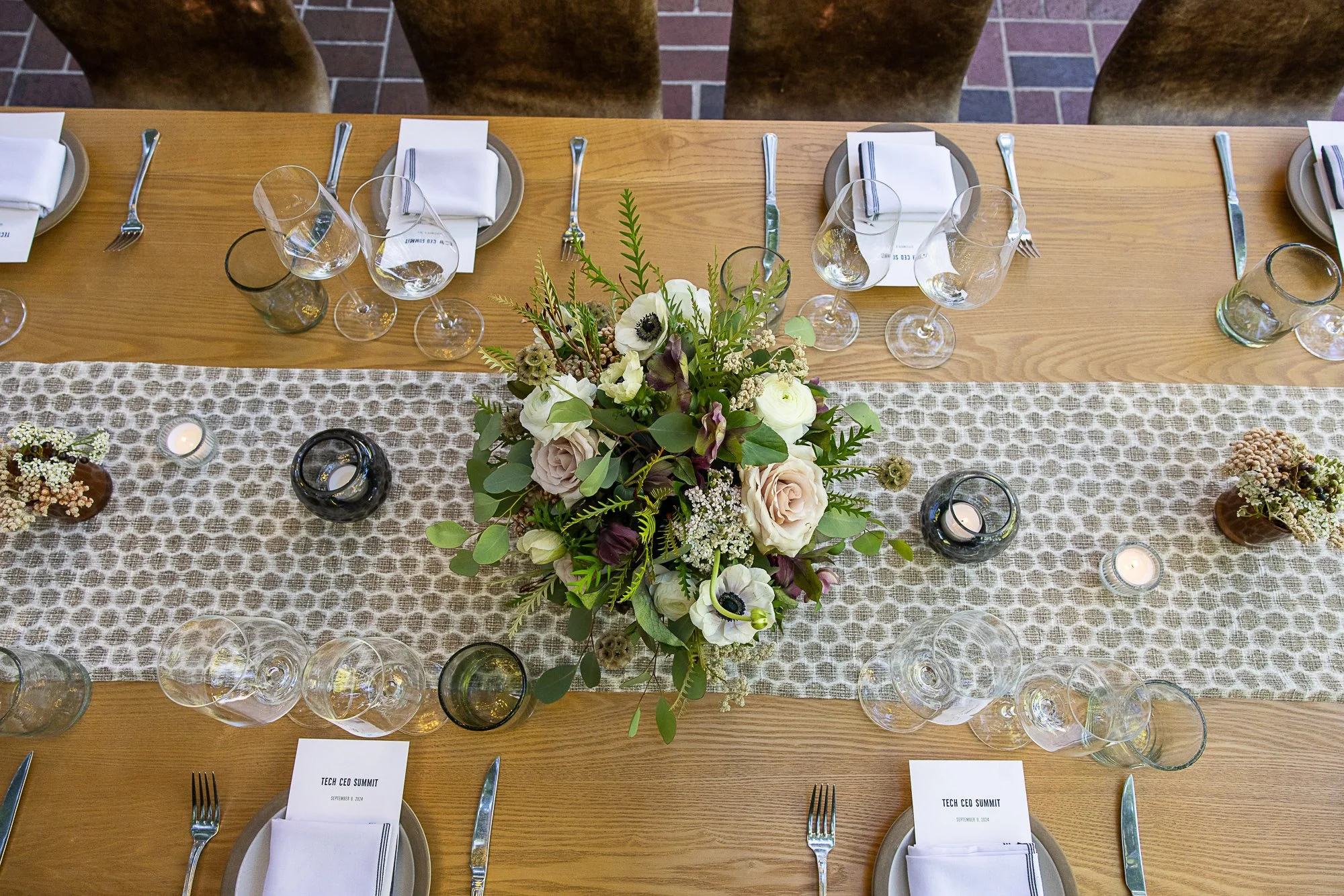 A long wooden table decorated with a floral centerpiece, candles, and place settings for a formal event labeled 'Tech CEO Summit' on September 8, 2024, with white napkins, plates, forks, knives, and empty glasses.