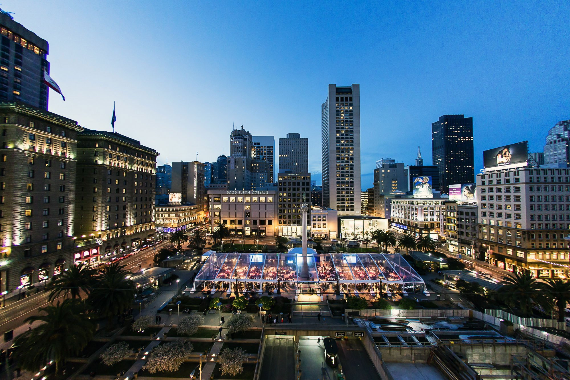 A cityscape of tall skyscrapers and buildings in the evening with illuminated windows and billboards, including one advertising Saks Fifth Avenue, and a glass rooftop shelter covering an outdoor seating area with people beneath.