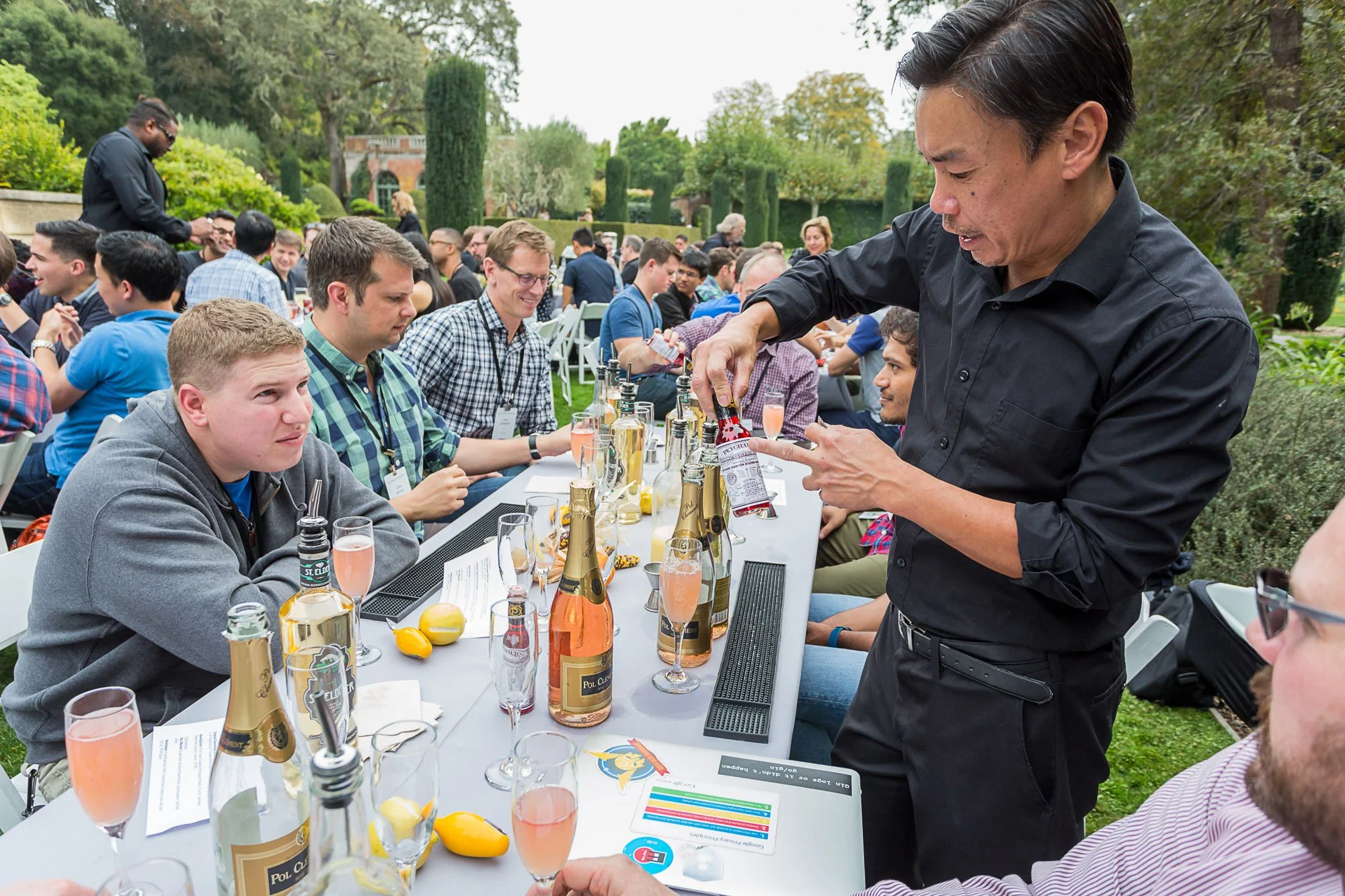 People attending an outdoor event or celebration, seated at a long table with bottles of rosé wine, lemons, and menus, with a man pouring drinks. Green garden with trees and shrubbery in the background.