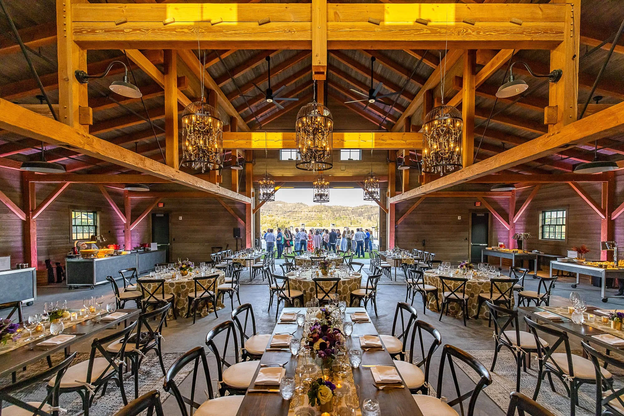 Interior view of a rustic banquet hall with wooden beams, chandeliers, and set tables ready for a wedding reception or event, with people gathering outside visible through open doors.