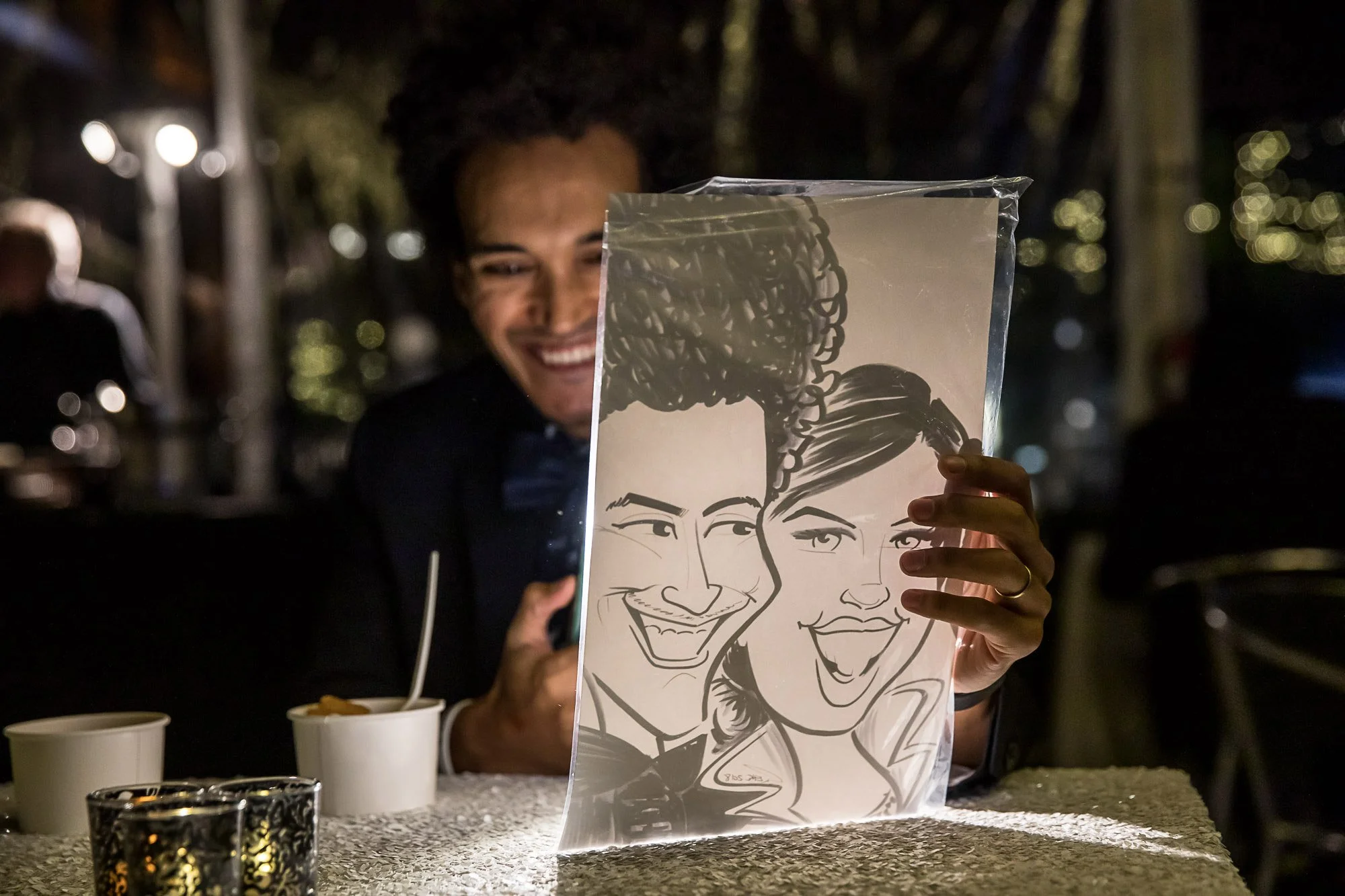 A person smiling and holding up a paper with a drawing of two smiling faces, in a nighttime outdoor setting with blurred lights and other people in the background.