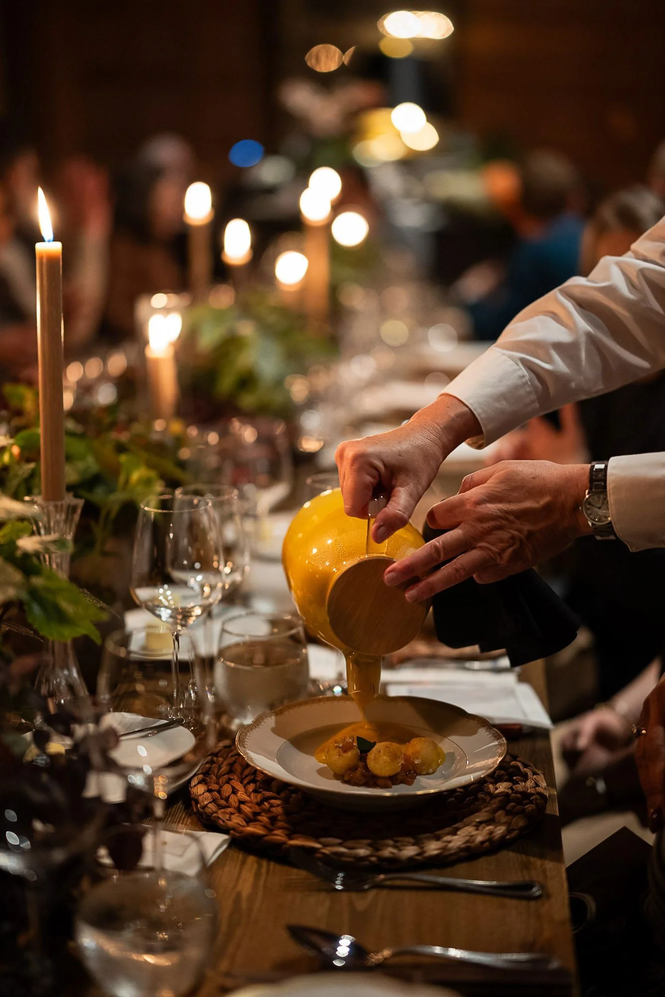 A person pouring sauce over a plated dish at a formal dinner with multiple guests, lit candles, and a decorated table.