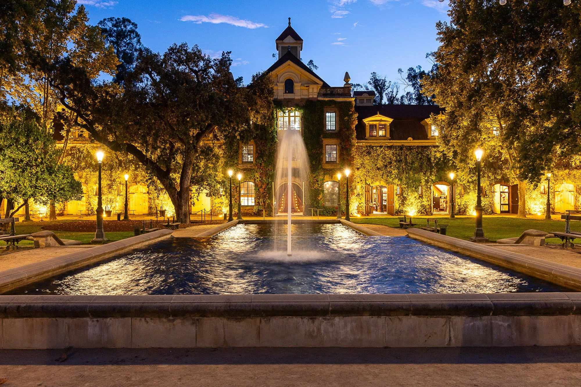 Nighttime view of a historic building with illuminated windows, surrounded by trees and vintage street lamps, featuring a water fountain in the foreground.