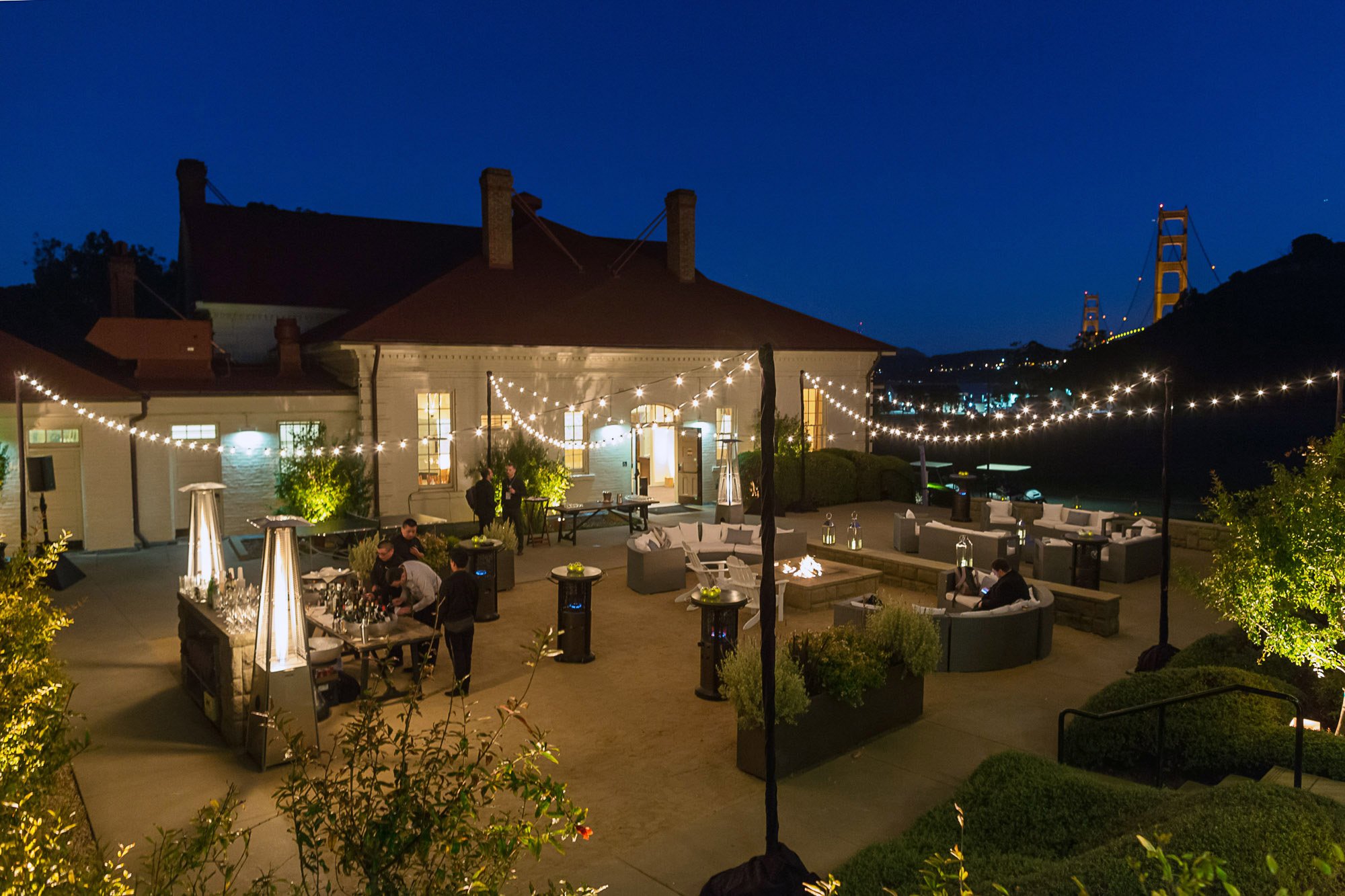 Nighttime outdoor gathering at a patio with string lights, seating, and a bar area, overlooking a bridge at night.