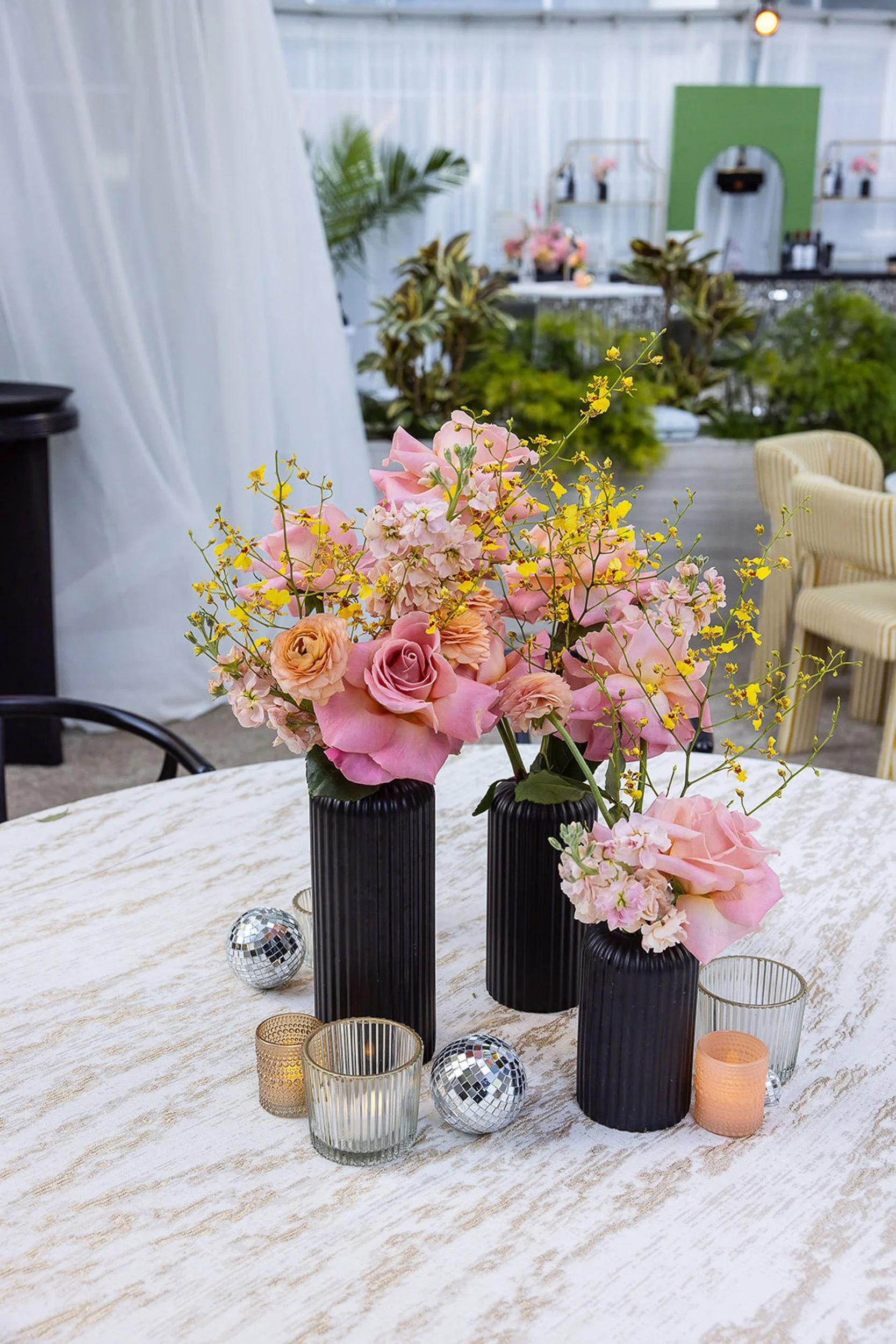 A table with floral centerpieces in black vases, small candle holders, and decorative objects, set in an event space with a white curtain backdrop and a bar area in the background.