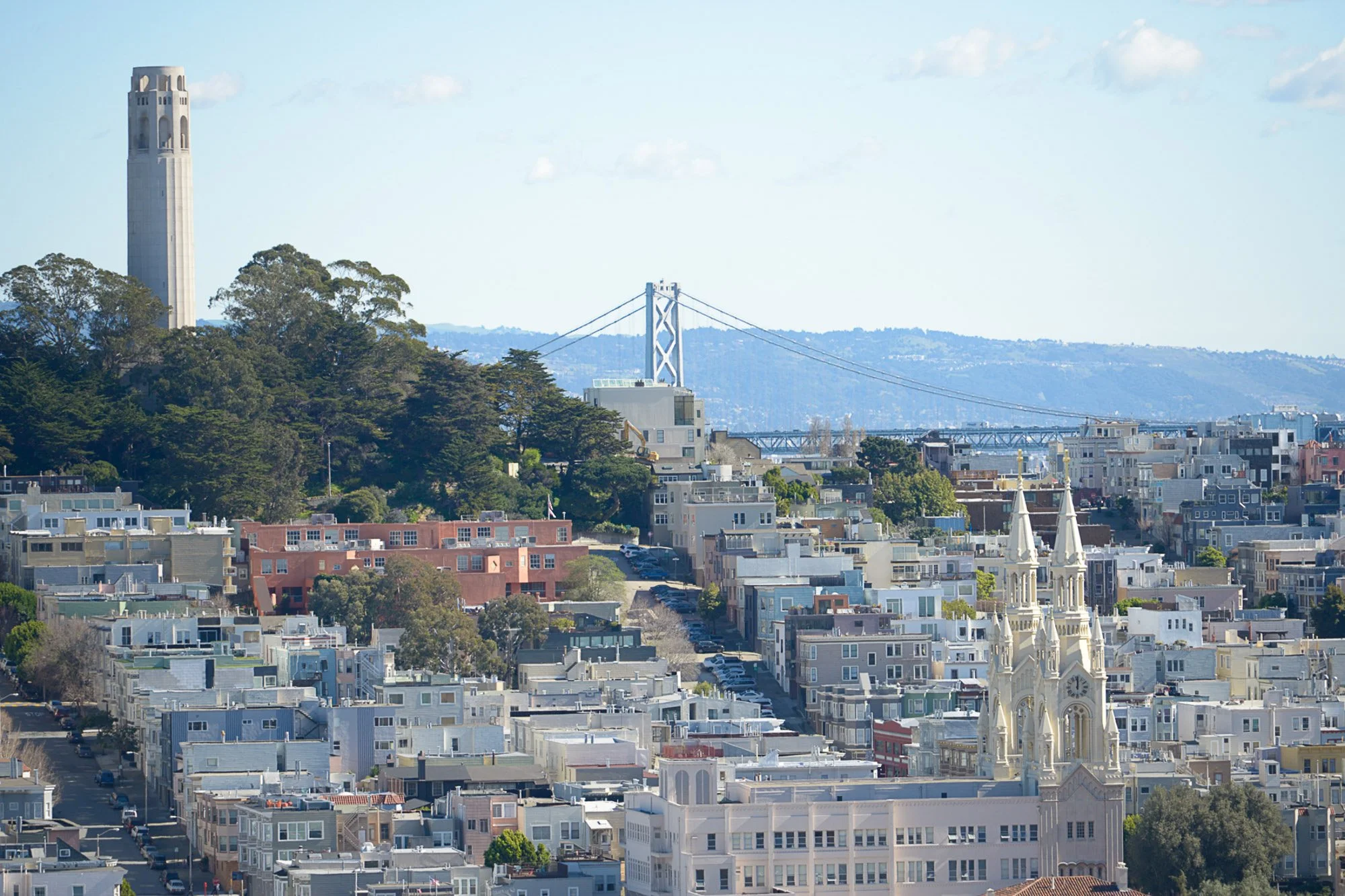 Cityscape of San Francisco featuring residential buildings, a church with white architecture, the Golden Gate Bridge in the background, and the Coit Tower on a hillside, under a partly cloudy sky.