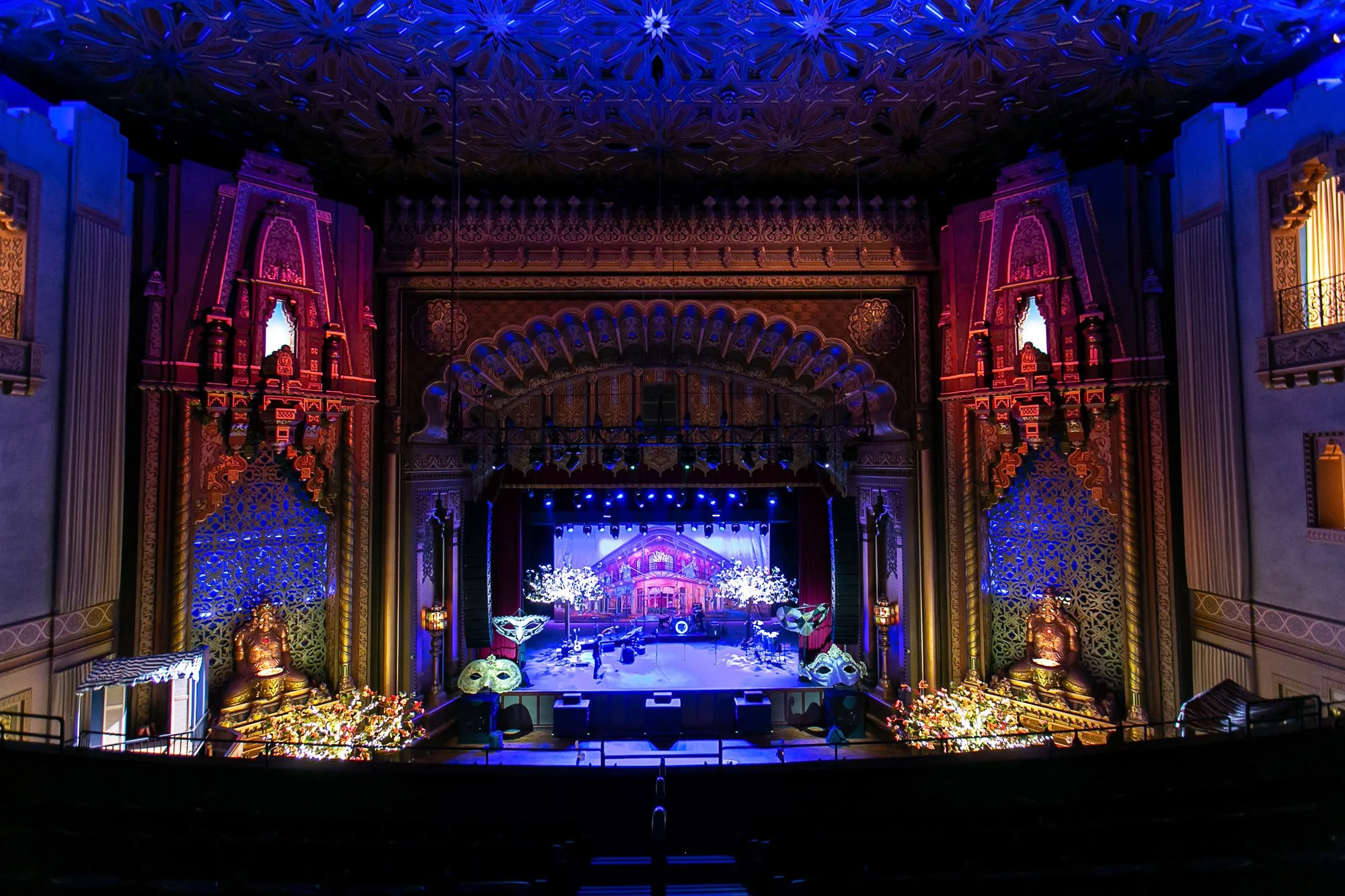 An ornate indoor stage decorated with bright, colorful lighting, with elaborate gold and purple architectural details, and floral arrangements in front.
