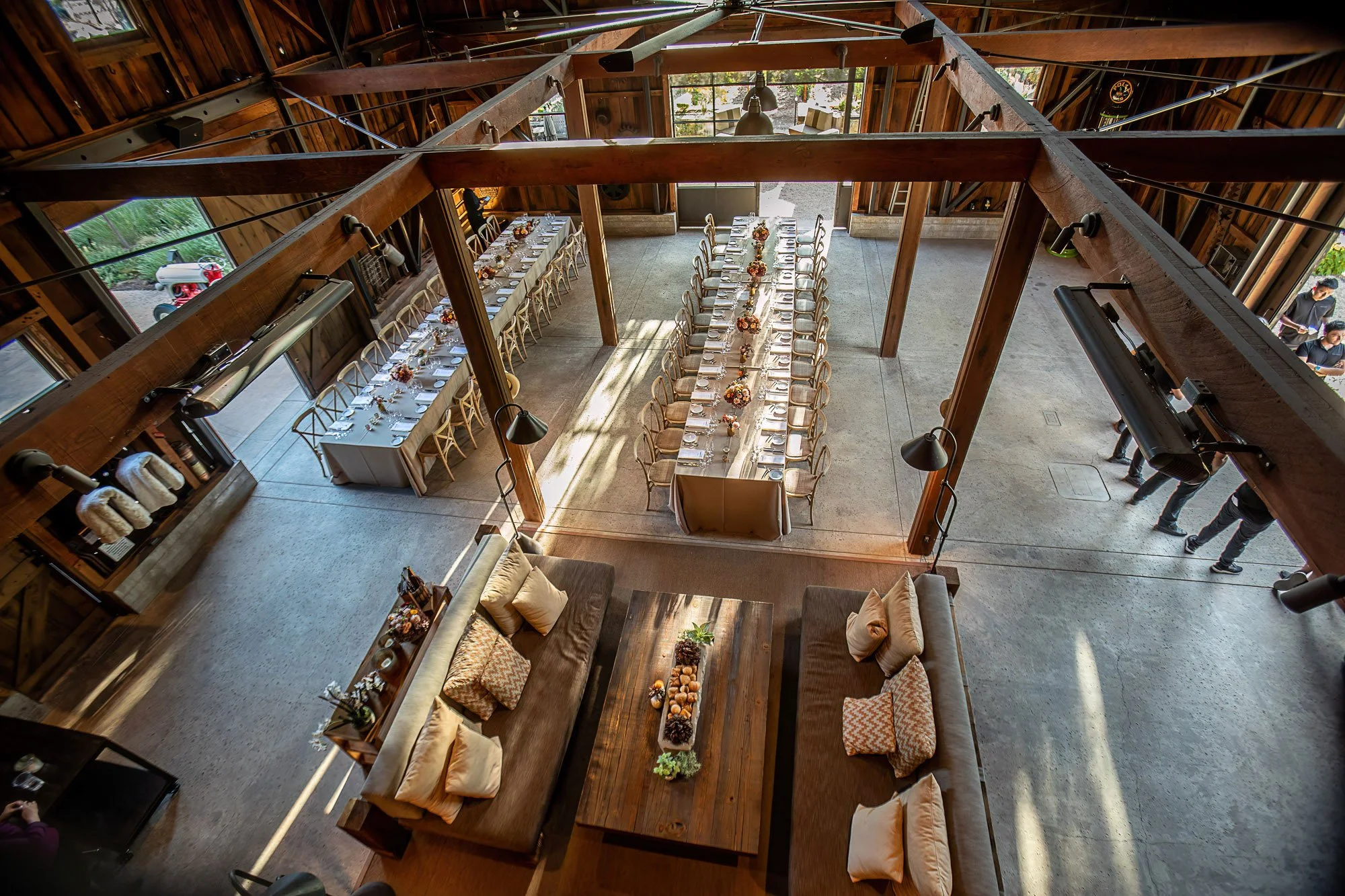 A rustic wooden barn interior set up for a wedding reception, with two long tables decorated with floral centerpieces, and seating for guests, featuring warm neutral tones and natural wood accents.