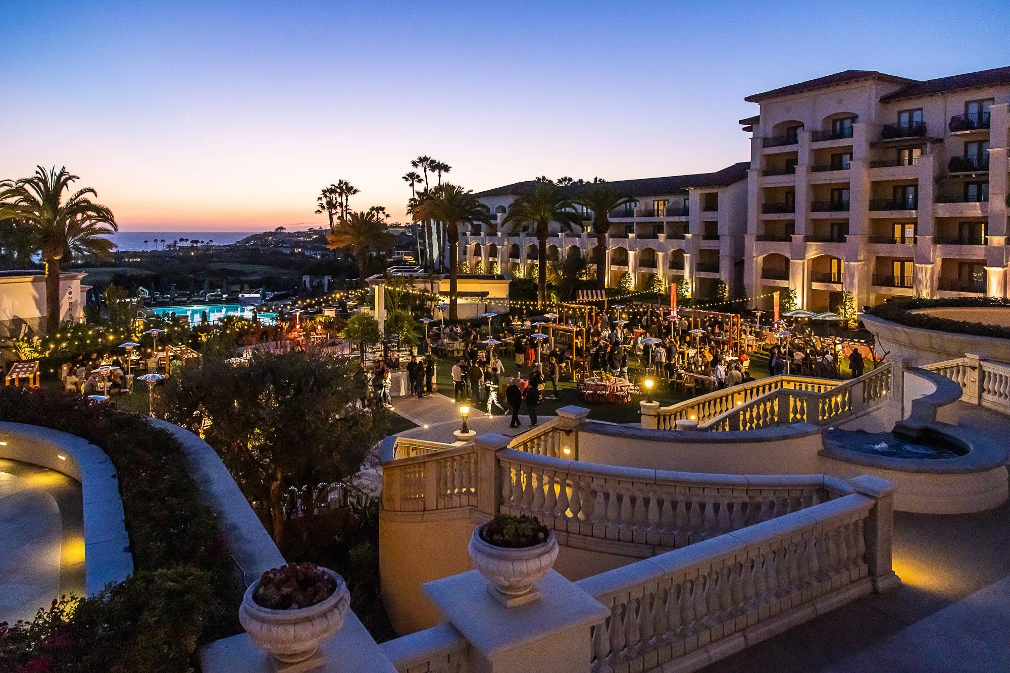 An evening scene at a resort with a crowd gathered on a patio, palm trees, a swimming pool, a large white hotel building, and ocean view in the background, with sunset sky.
