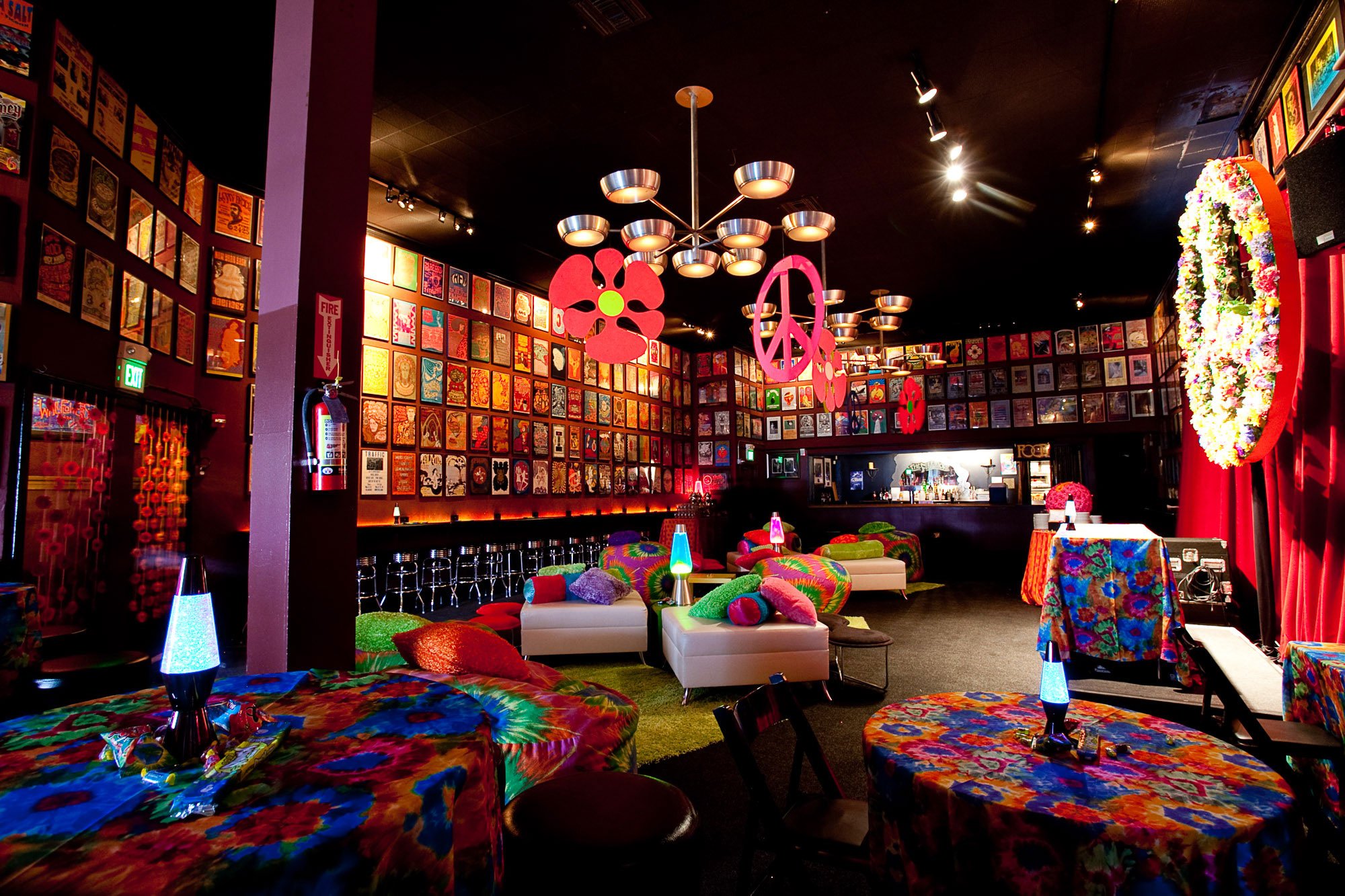 Colorful lounge area with psychedelic tables, plush pillows, vibrant chairs, flower and peace sign decorations, murals, and dim lighting in a retro-style space.