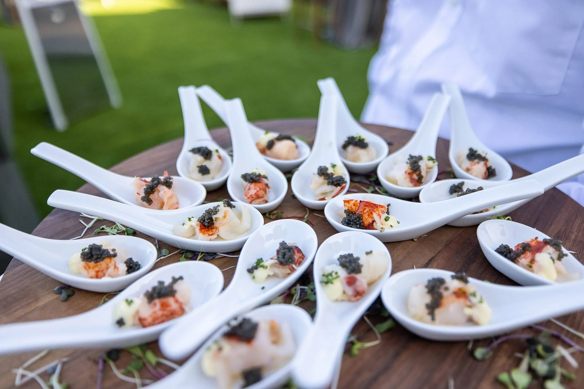Multiple white ceramic spoons arranged in a semi-circle on a wooden table, each containing a gourmet appetizer with seafood, garnished with black caviar and herbs. The background shows a blurred outdoor setting with green grass.