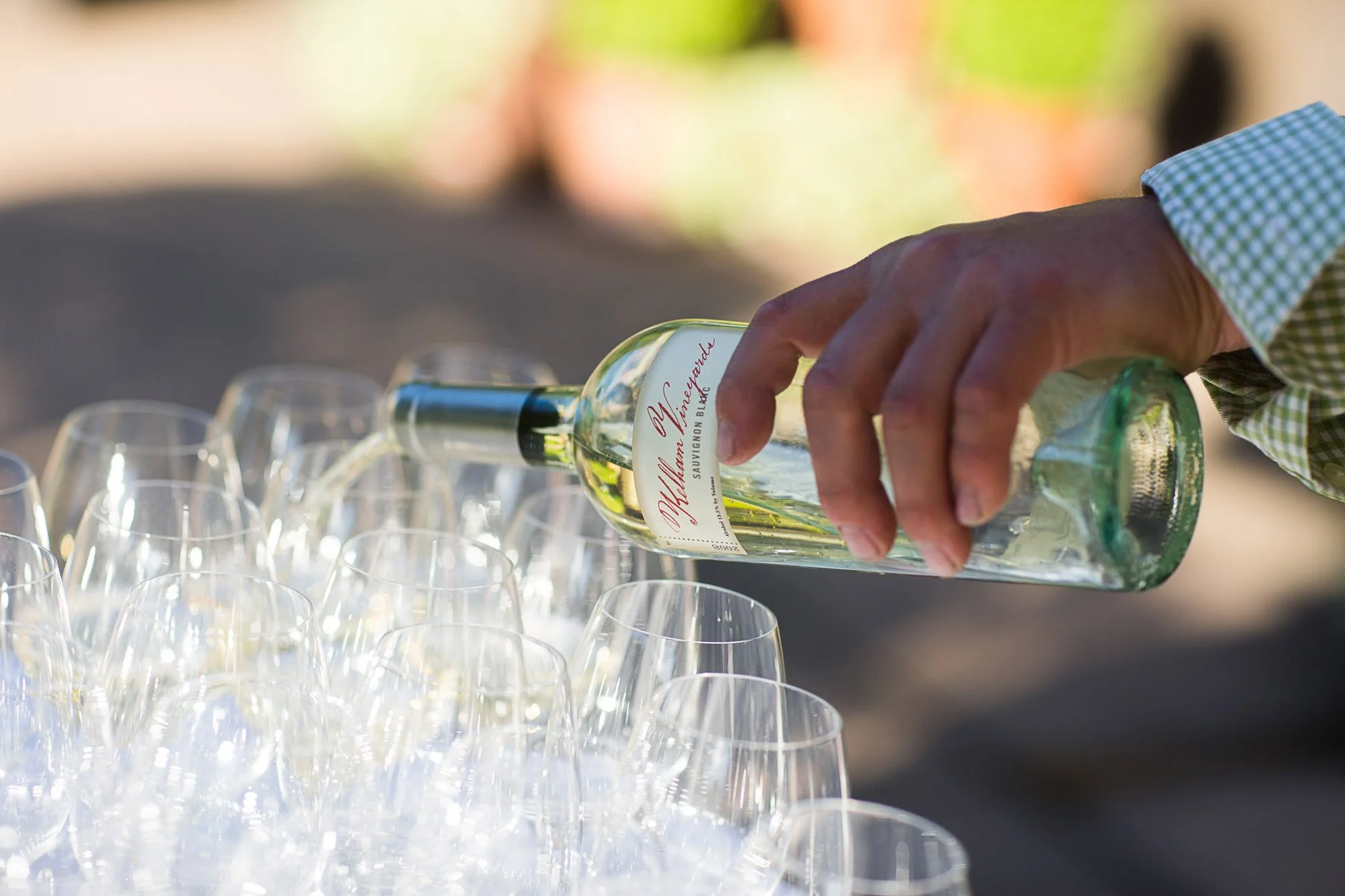 A hand pouring white wine from a bottle into multiple empty wine glasses on a table outdoors.