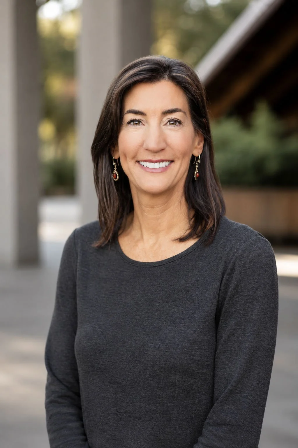 A woman with medium-length dark brown hair, smiling, wearing a dark gray long-sleeve top and red earrings, standing outdoors with blurred background of trees and a structure.
