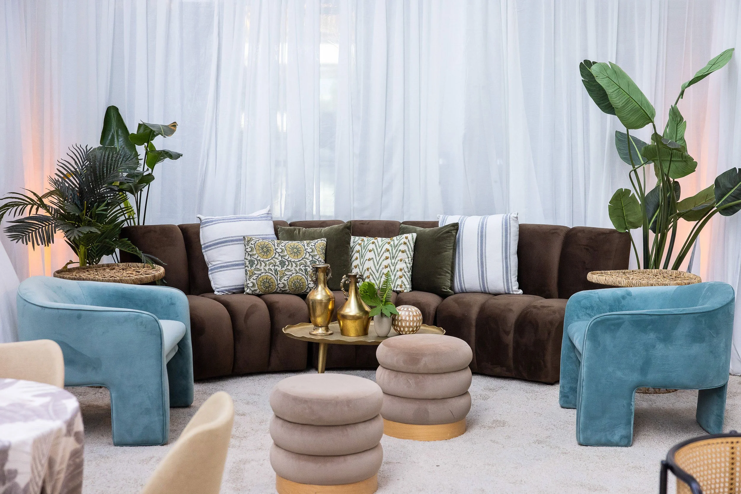 Living room with brown sofa and blue armchairs, decorated with patterned and solid cushions, large green plants on both sides, white curtains in the background, and a small round beige ottoman in front.