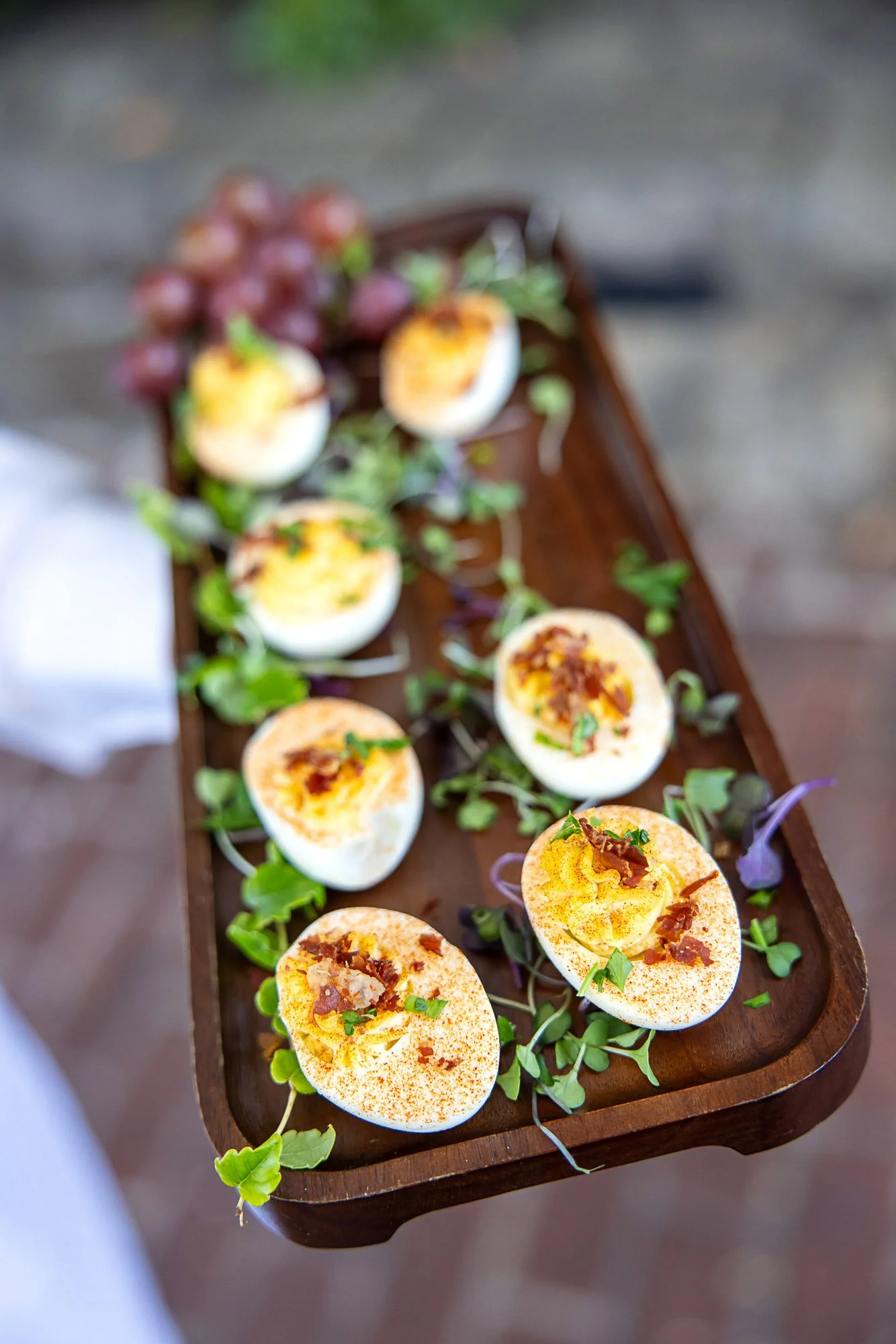 A wooden platter with sliced hard-boiled eggs garnished with herbs and bacon bits, surrounded by microgreens on a blurred wooden surface.