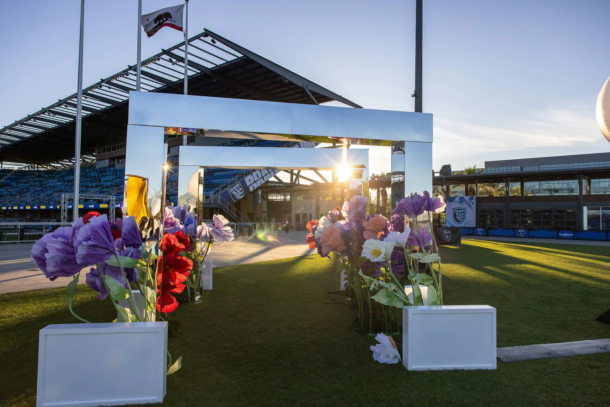 The image shows a sports stadium entrance with a large rectangular mirror frame, decorated with colorful flowers at its base. In the background, there is a stadium seating area, a flagpole with the California state flag, and a modern building. The su