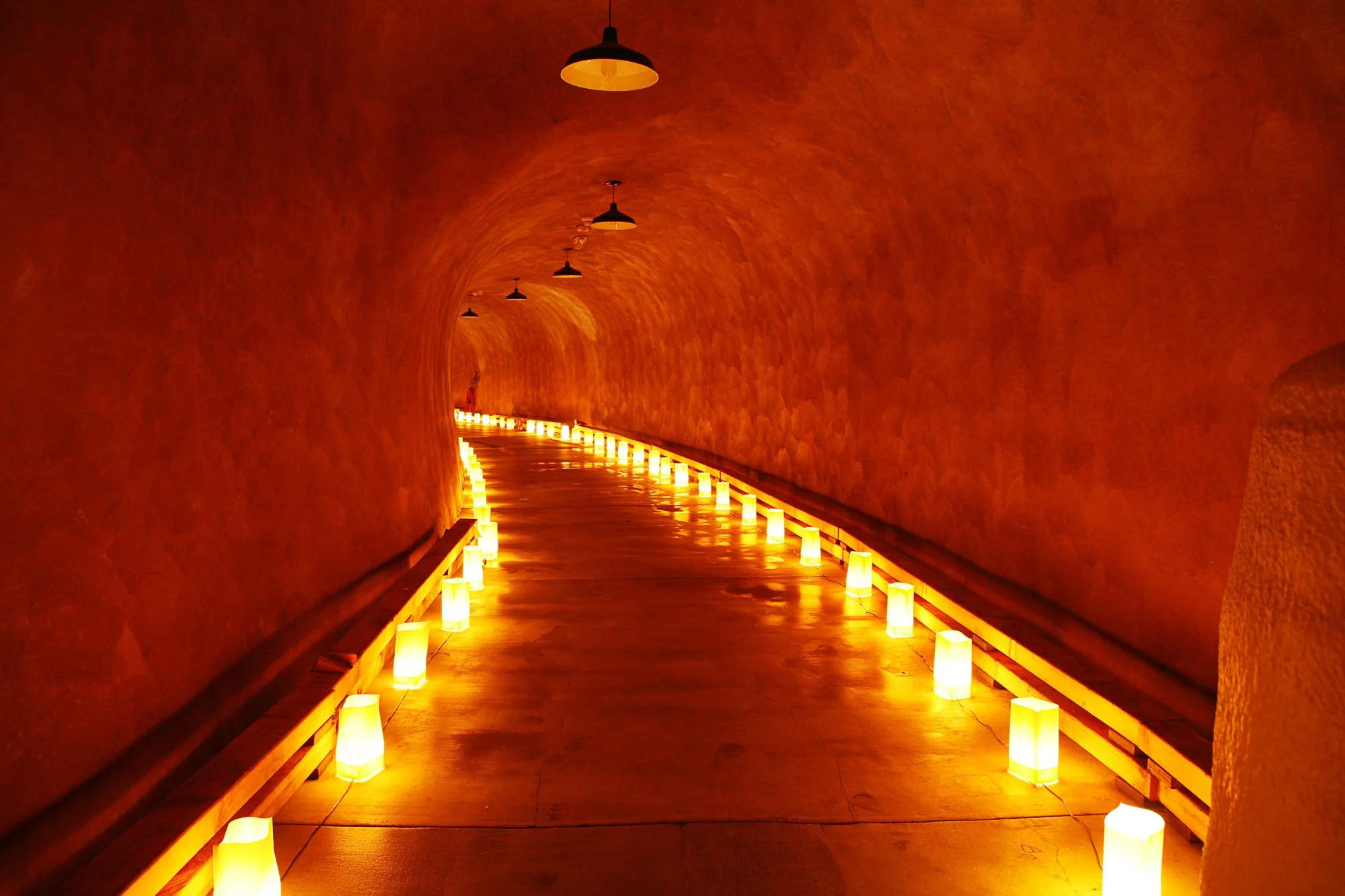 A tunnel with warm-colored wall lights along the pathway, hanging pendant lights on the ceiling, and a curved, enclosed brick wall design.