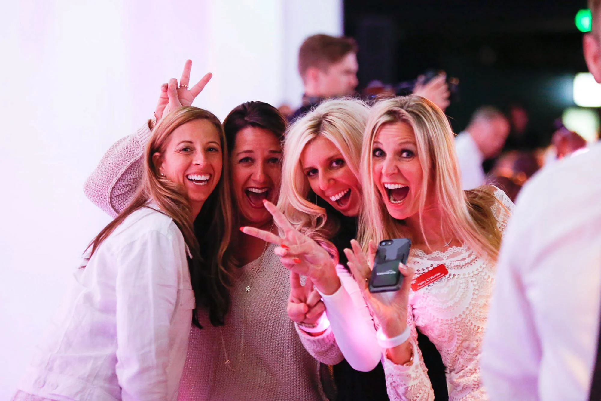 Four women taking a selfie at a social event, with a man in the background holding a camera.