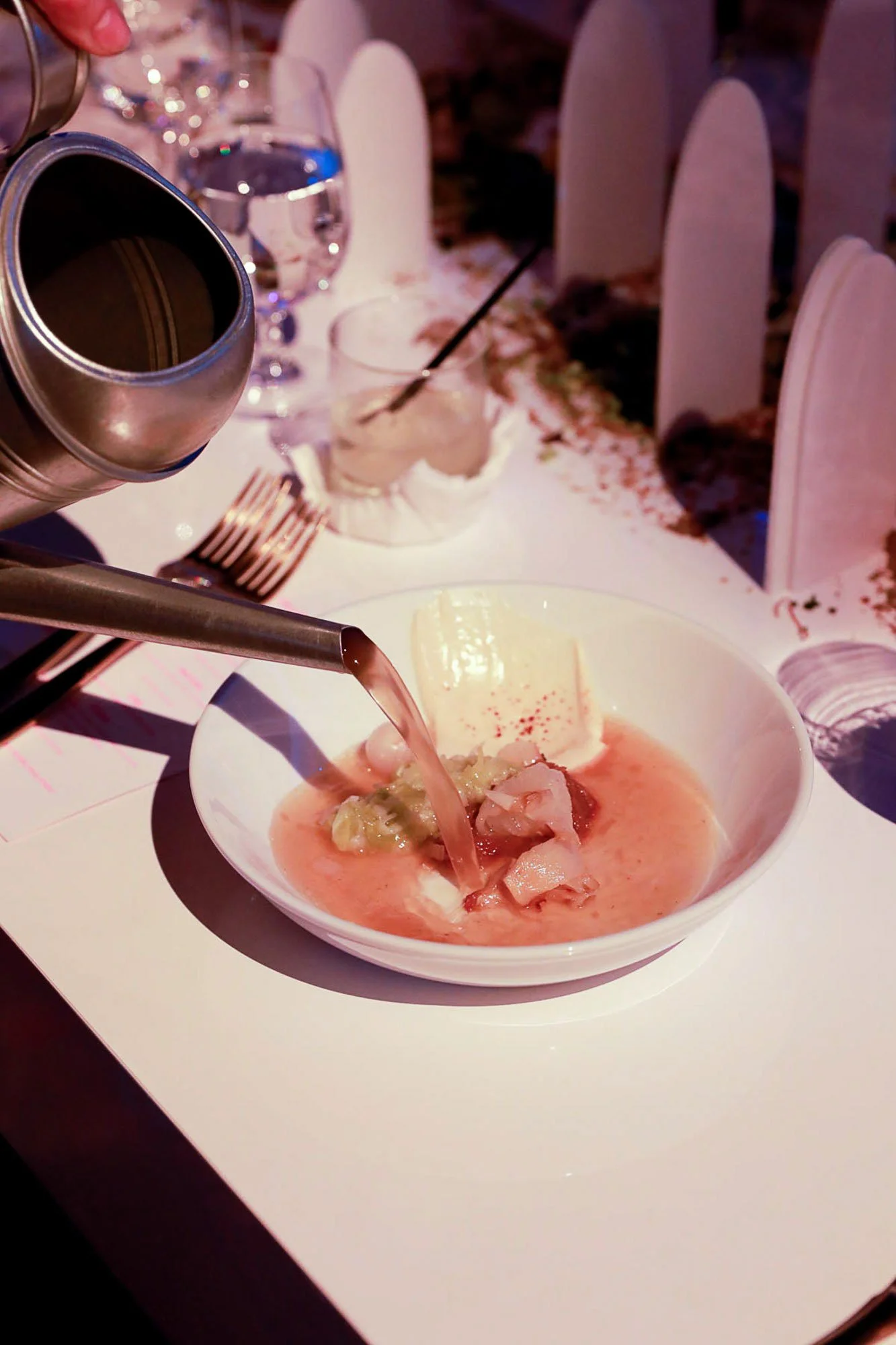 A bowl of pink-colored seafood soup with chunks of fish and vegetables, on a white table, with a glass of water, a fork, and a layered dessert in the background.