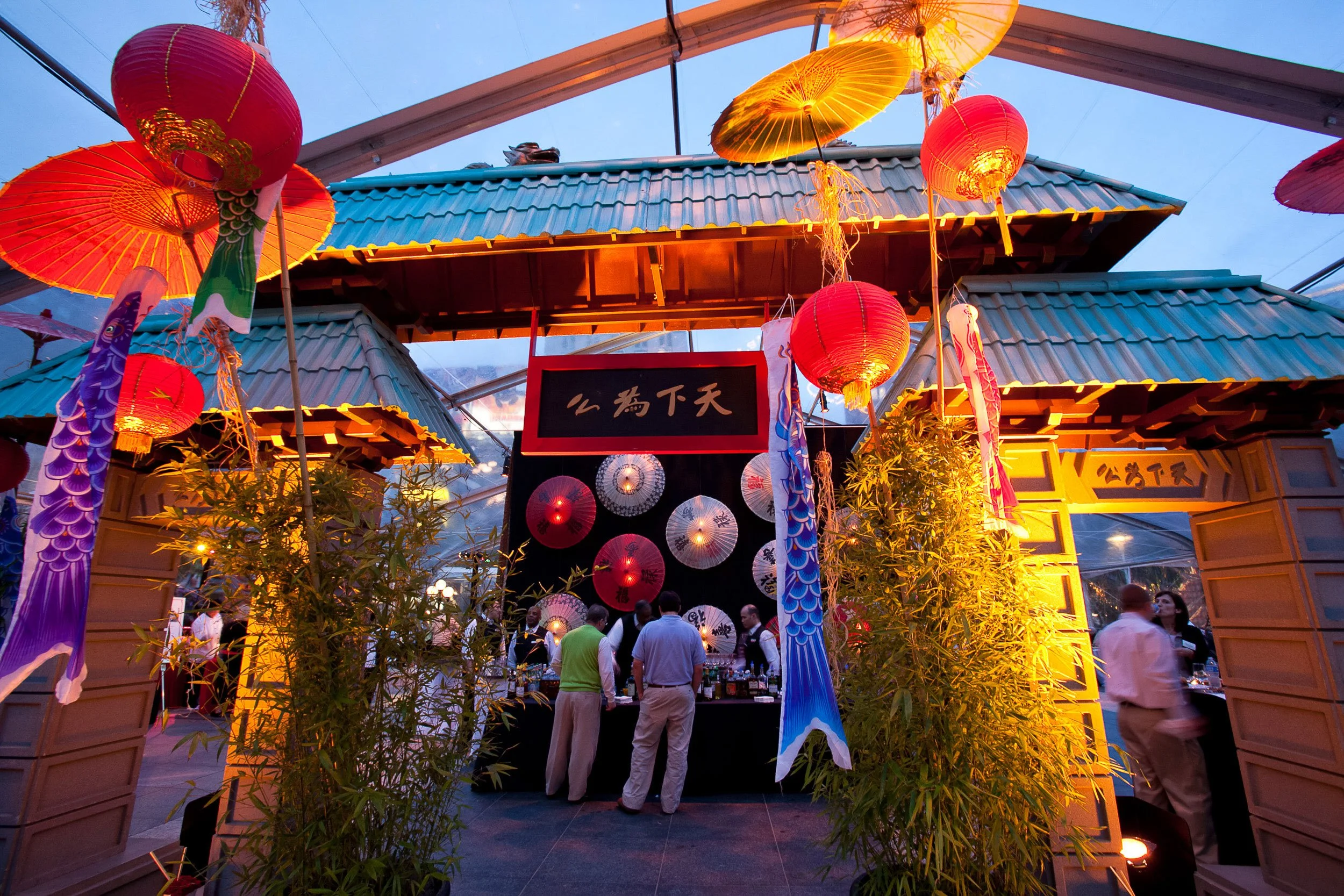 Night scene at an outdoor Asian festival with colorful hanging lanterns, umbrellas, and a crowd of people.