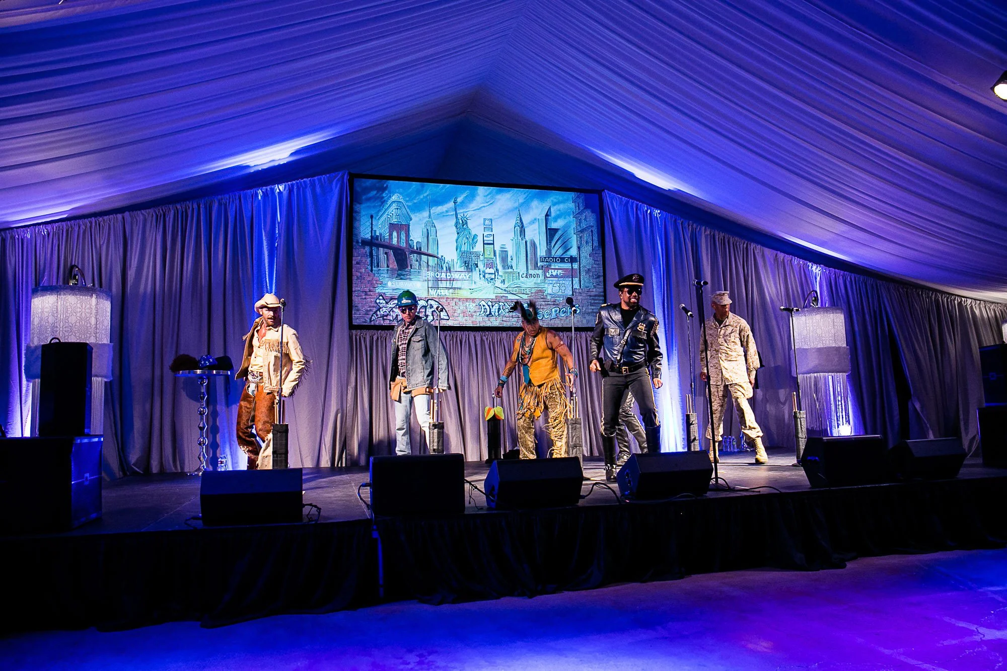 Group of performers dressed in various costumes on stage, with a backdrop displaying New York City landmarks, under purple lighting.