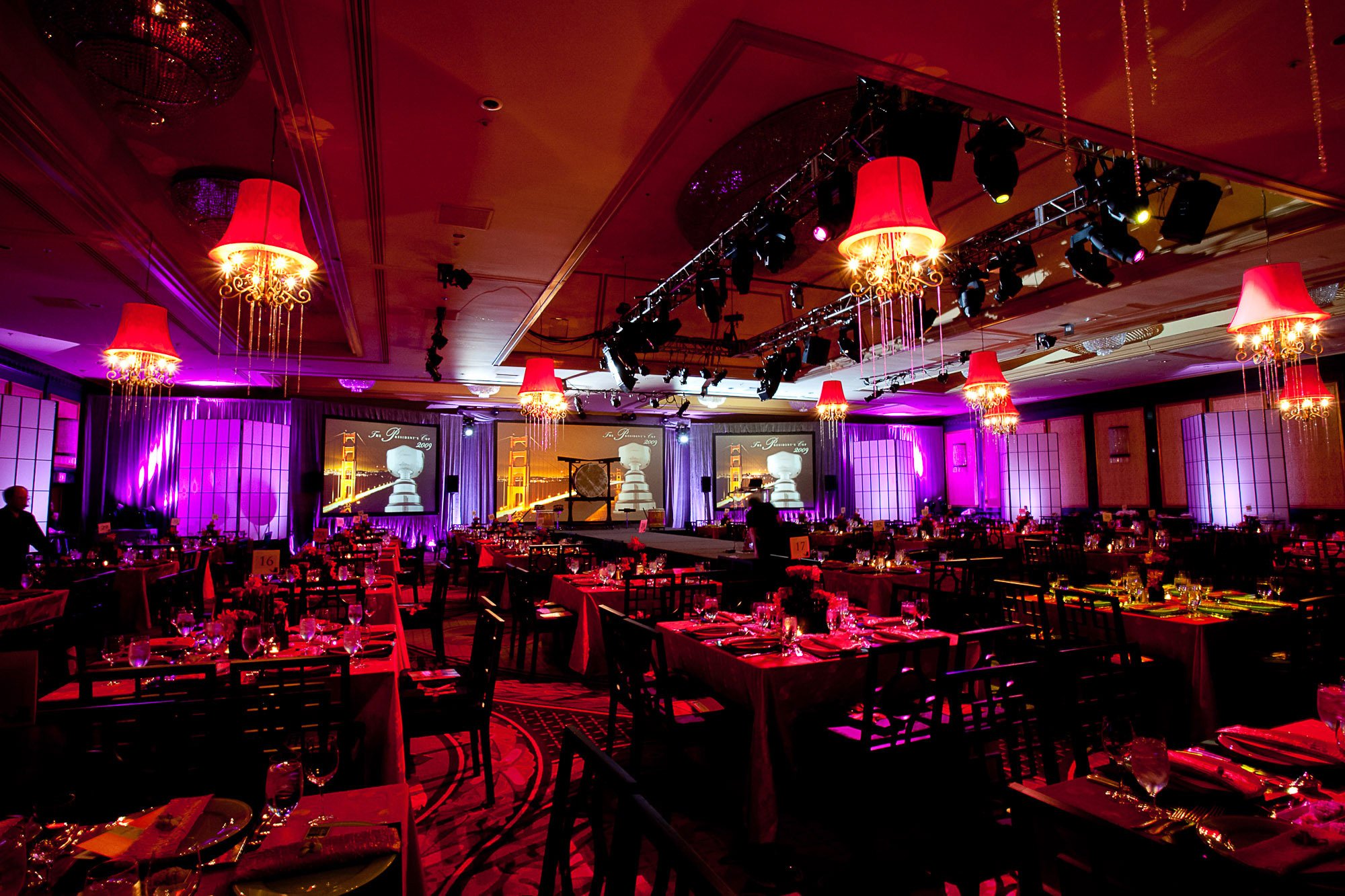 An elegant banquet hall decorated with red lampshade chandeliers, purple lighting, and multiple round tables set with glassware and napkins, prepared for a formal event or celebration.