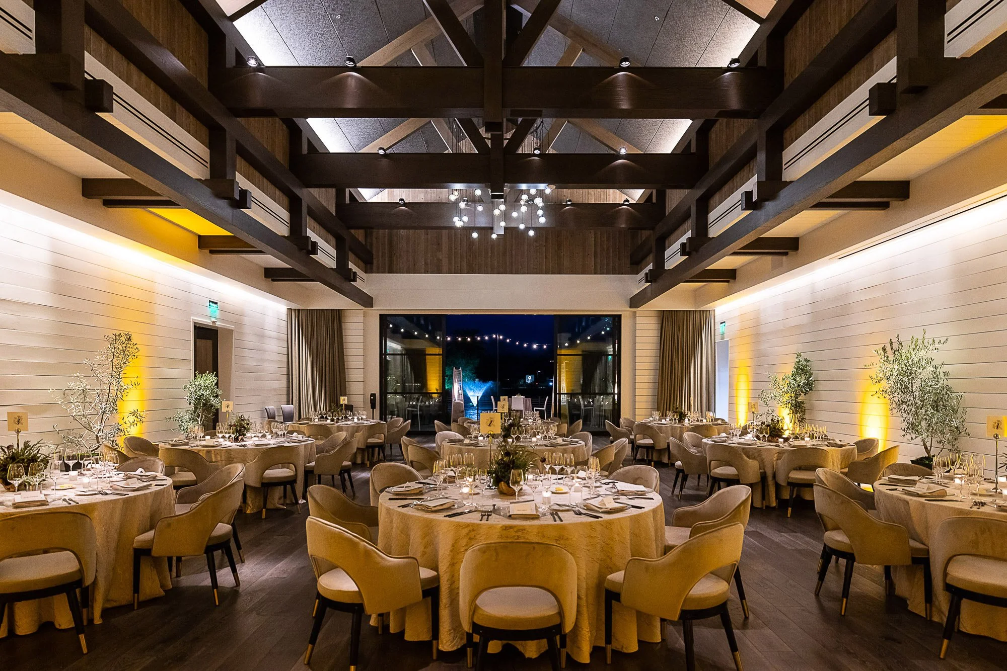 Elegant event space with round tables draped in light-colored tablecloths, set with glassware and centerpieces, surrounded by beige chairs; high ceiling featuring dark wood beams and modern chandelier; walls with soft lighting and large windows with 