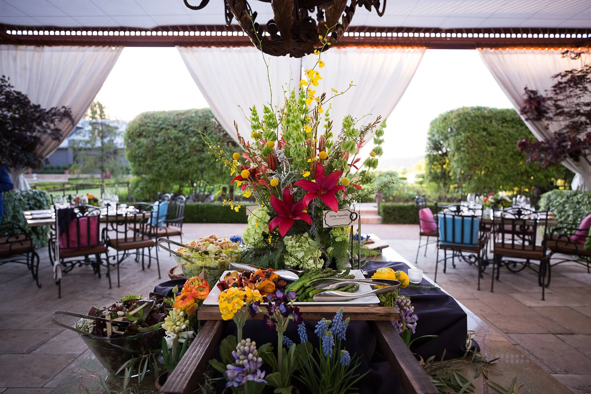 A table with a floral centerpiece and assorted dishes in an outdoor or semi-outdoor setting, with chairs and a view of greenery through large open windows or curtains.