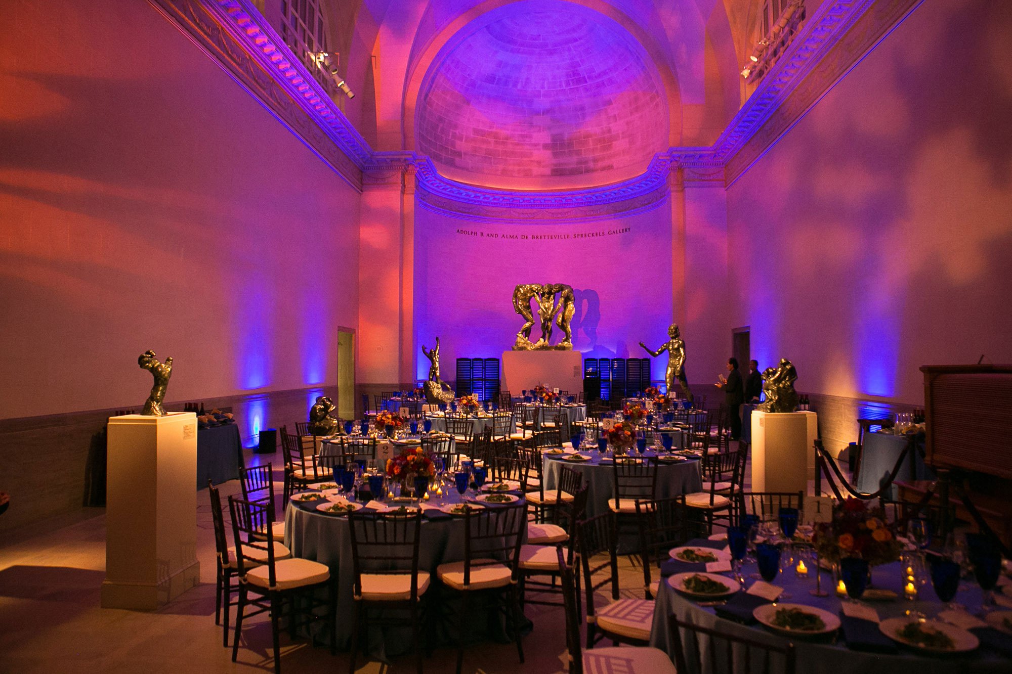 Elegant banquet hall decorated with colorful lighting, round tables set with blue glasses, floral centerpieces, and gold sculptures of human figures, with a large curved ceiling and a sculpture display at the back.