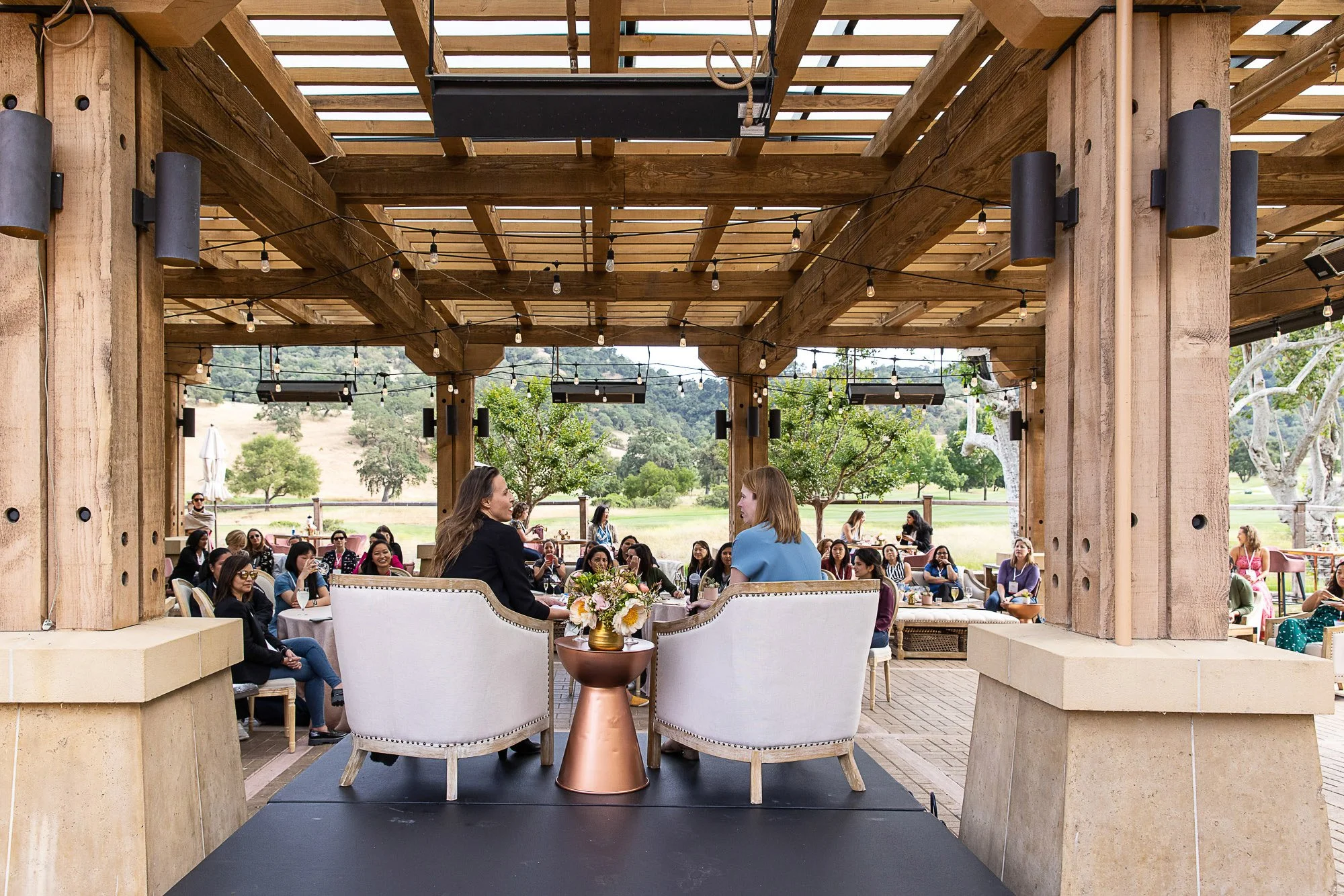 People gathered at an outdoor event under a wooden pergola with string lights, seated at tables with floral centerpieces, with a scenic landscape of trees and hills in the background.
