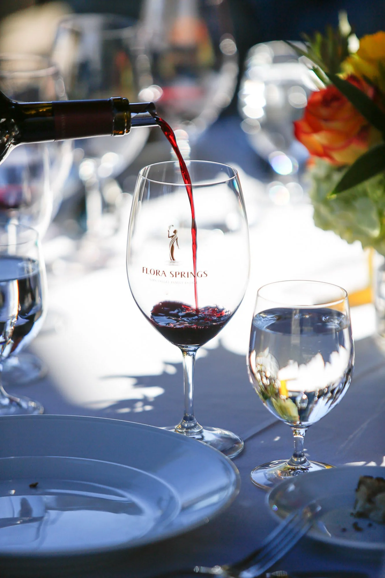 Red wine being poured from a bottle into a wine glass labeled Flora Springs. Other glasses and plates are on the table, with a blurred floral arrangement on the side.