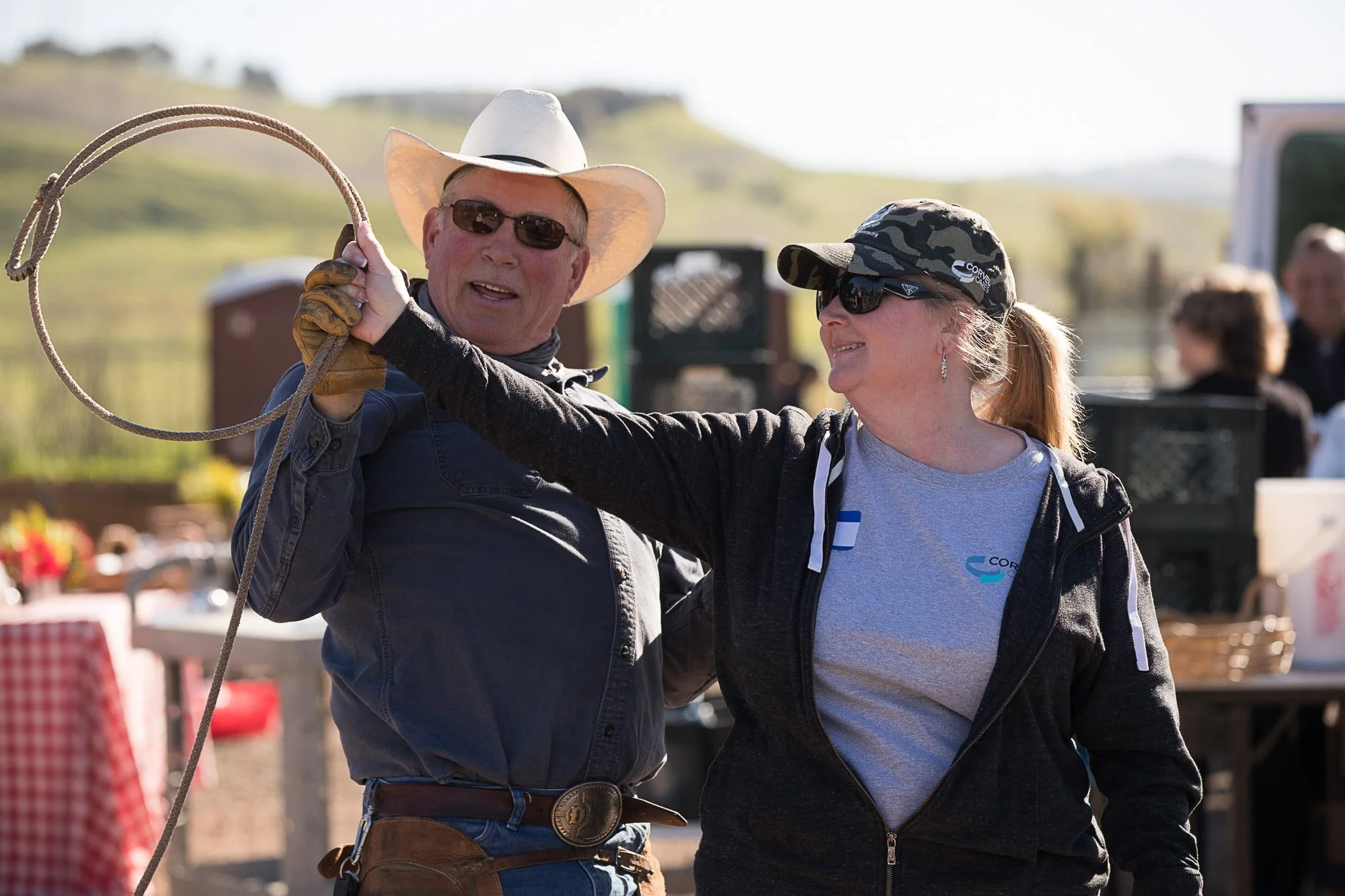 Two people, a man and a woman, are engaged in an outdoor activity involving a lasso, at a fair or outdoor event. The man is wearing a white cowboy hat, sunglasses, and a dark jacket. The woman has her hair in a ponytail, is wearing sunglasses, a camo