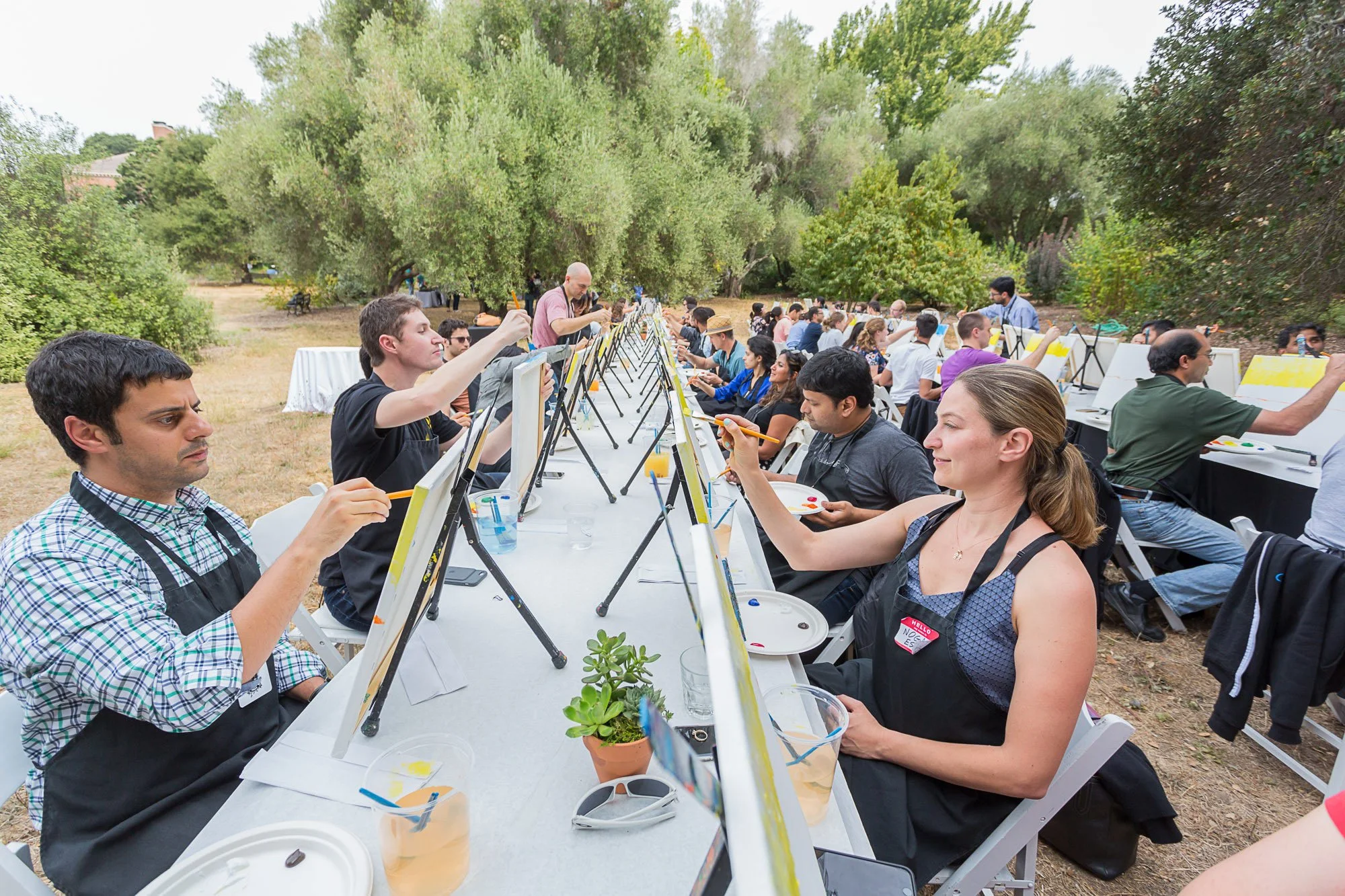 Group of people participating in outdoor painting event, seated at long tables with easels, surrounded by trees.