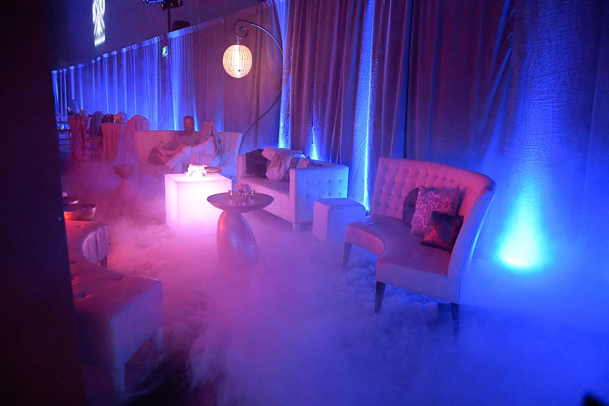 A dimly lit lounge with blue and purple lighting, featuring plush white sofas with pillows, a small round table, and a decorative hanging lamp. There are two people sitting on a sofa, engaged in conversation, and the atmosphere appears relaxed and co