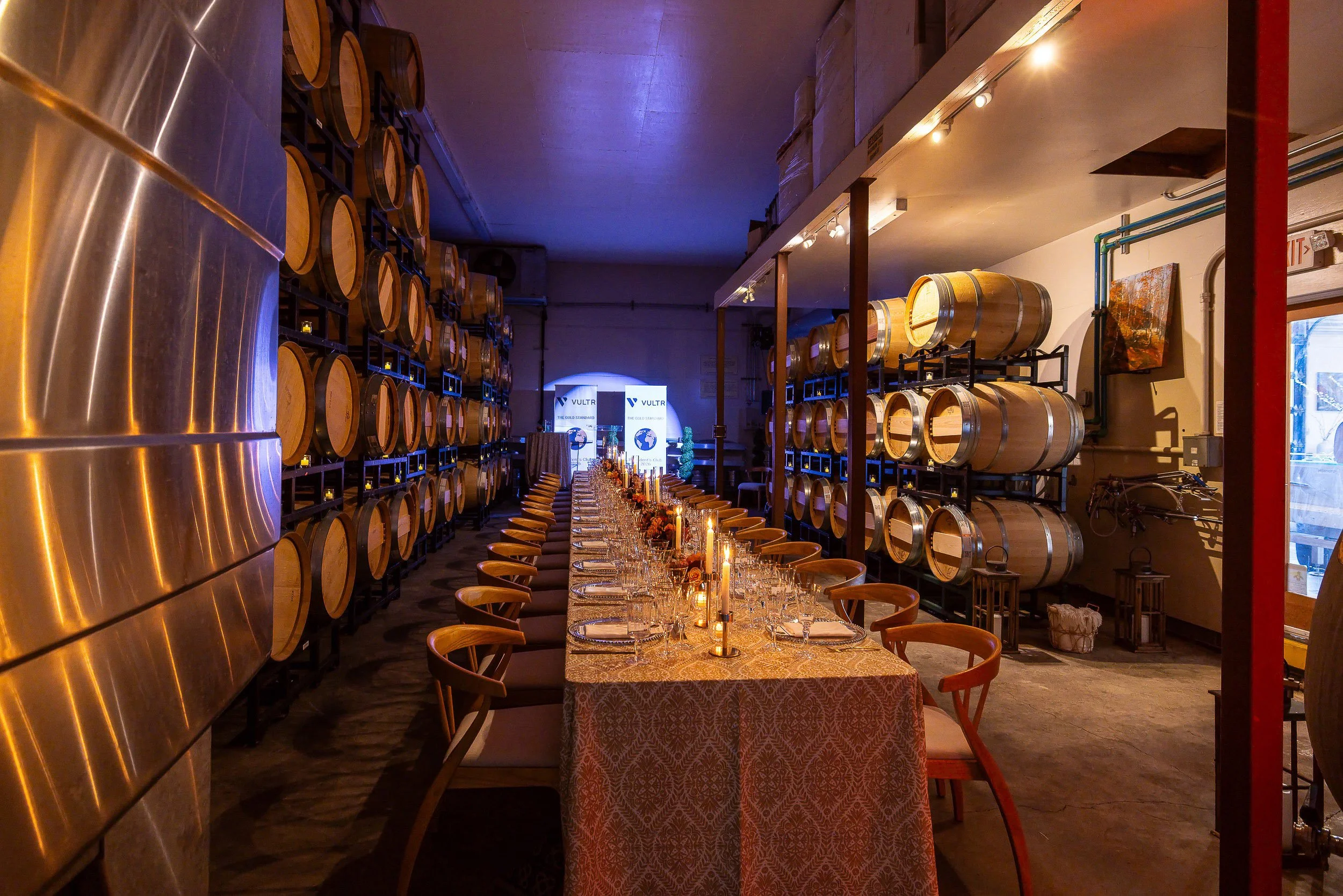 Long dining table with chairs set for a meal, surrounded by wine barrels stored on metal racks in a rustic restaurant or wine cellar, illuminated with warm lighting and candles.