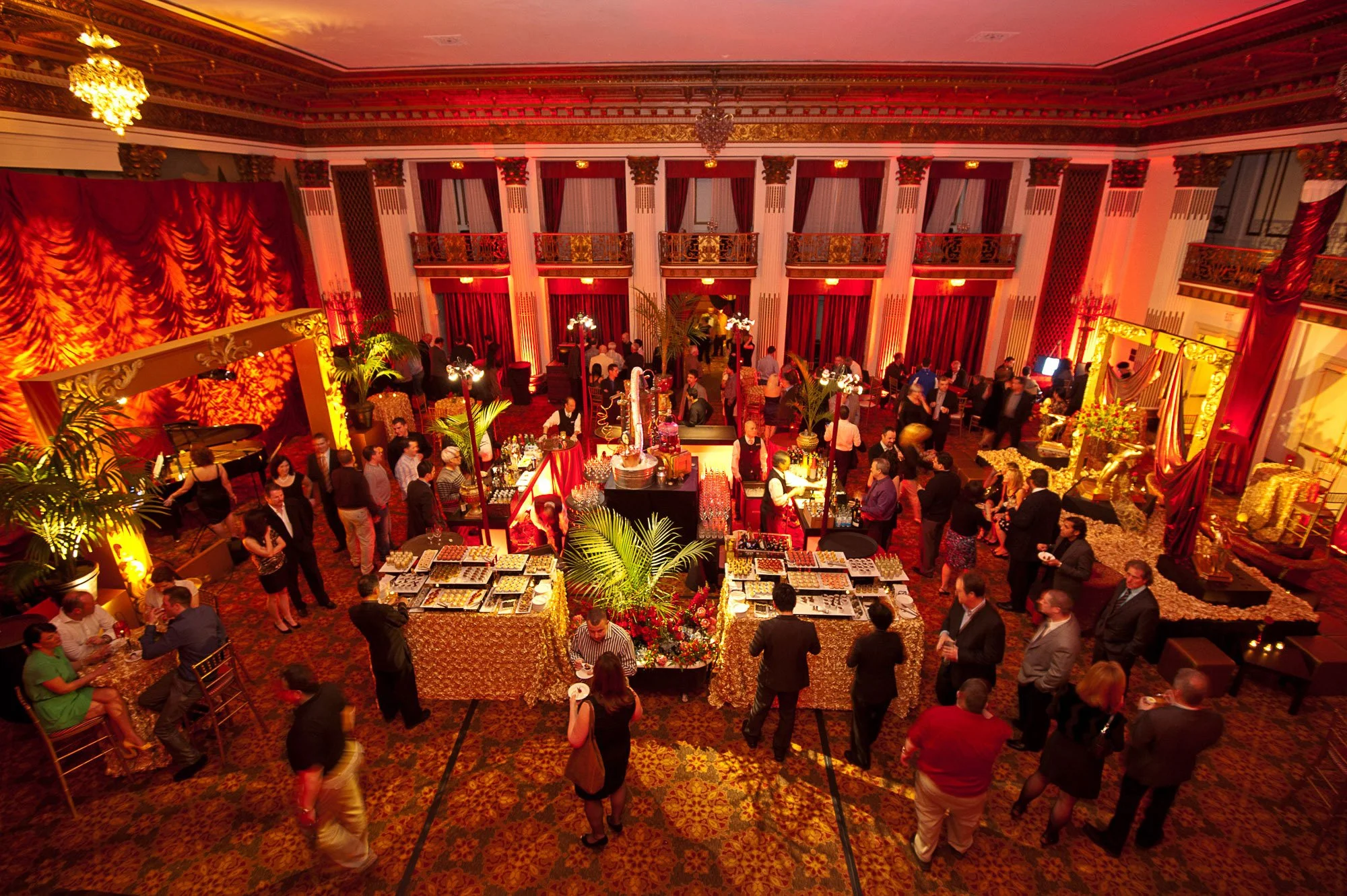 An elegant ballroom filled with people attending a social event. The room has gold and red decor, with large red curtains and ornate gold accents. There are tables with food and drinks, and a bar area in the foreground. People are mingling, and the s