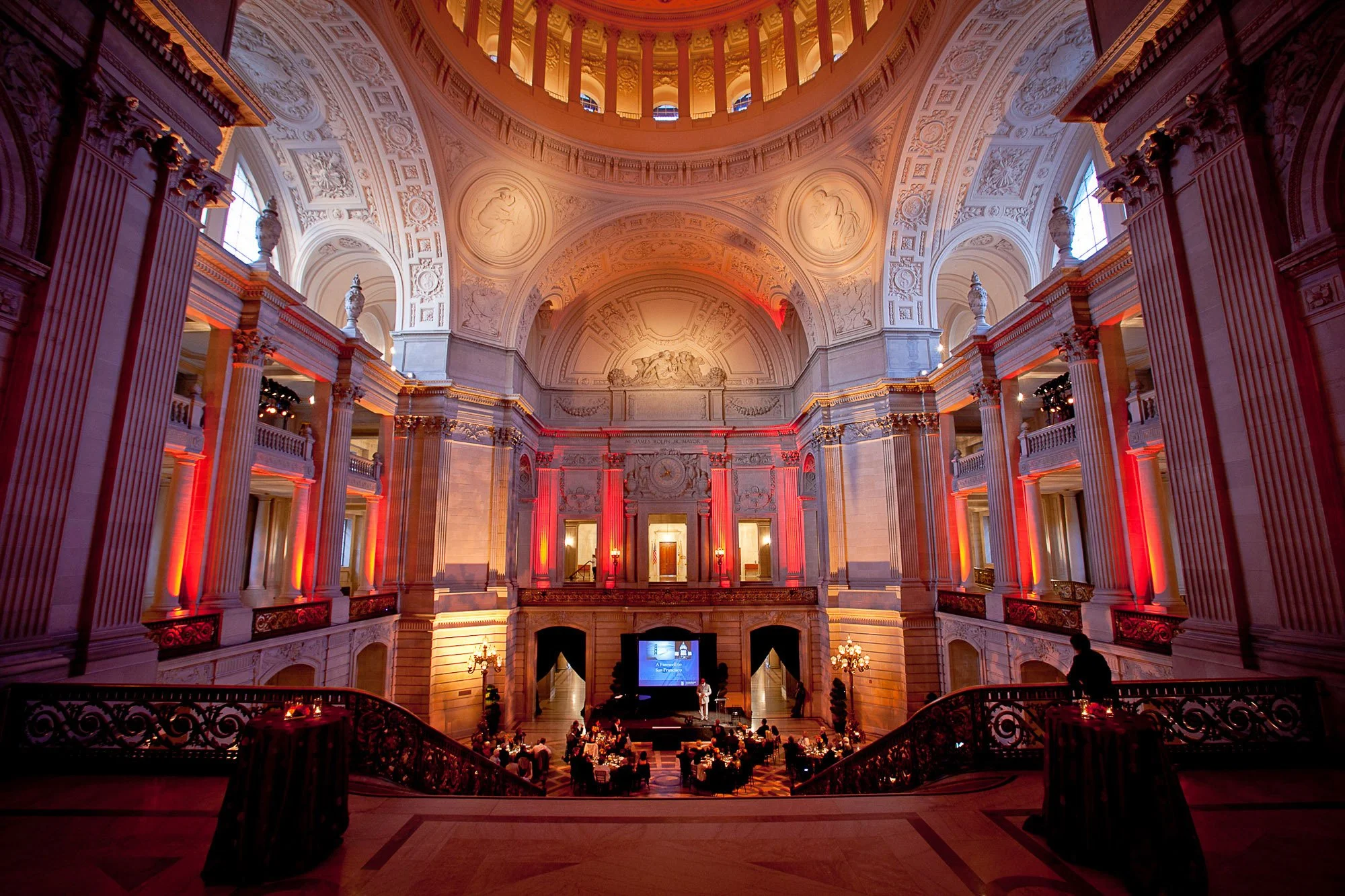 Interior of a grand historic building, possibly a theater or concert hall, featuring ornate architecture with decorative relief sculptures, columns, and balconies. Indoors, there is a stage with a presentation monitor, and the room is illuminated wit