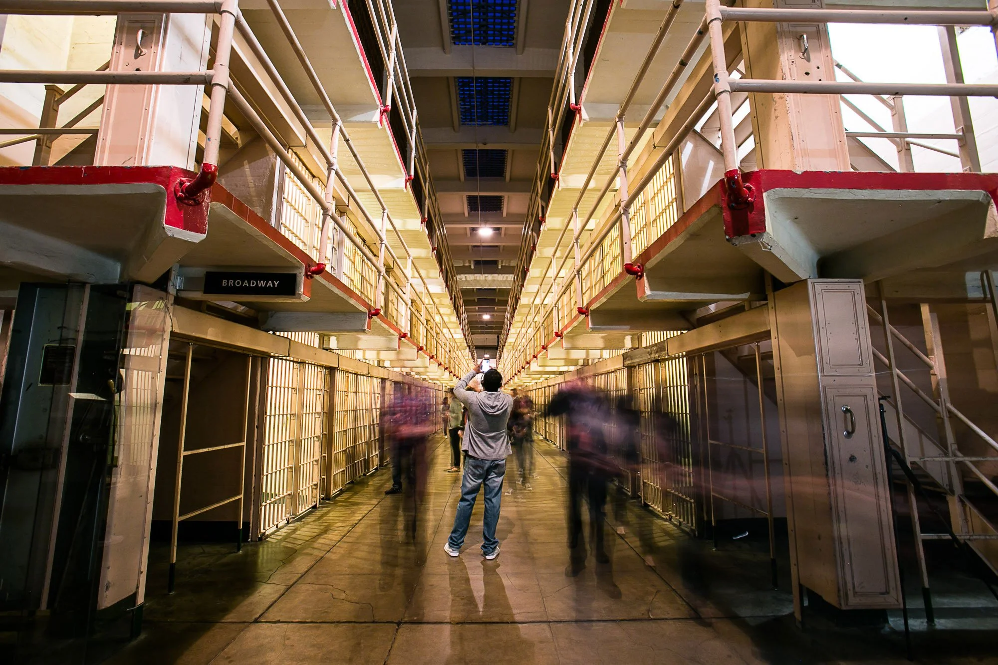 Interior of a building with metal scaffolding, yellow walls, and multiple people, some blurred from motion, walking through a corridor labeled 'Broadway'.