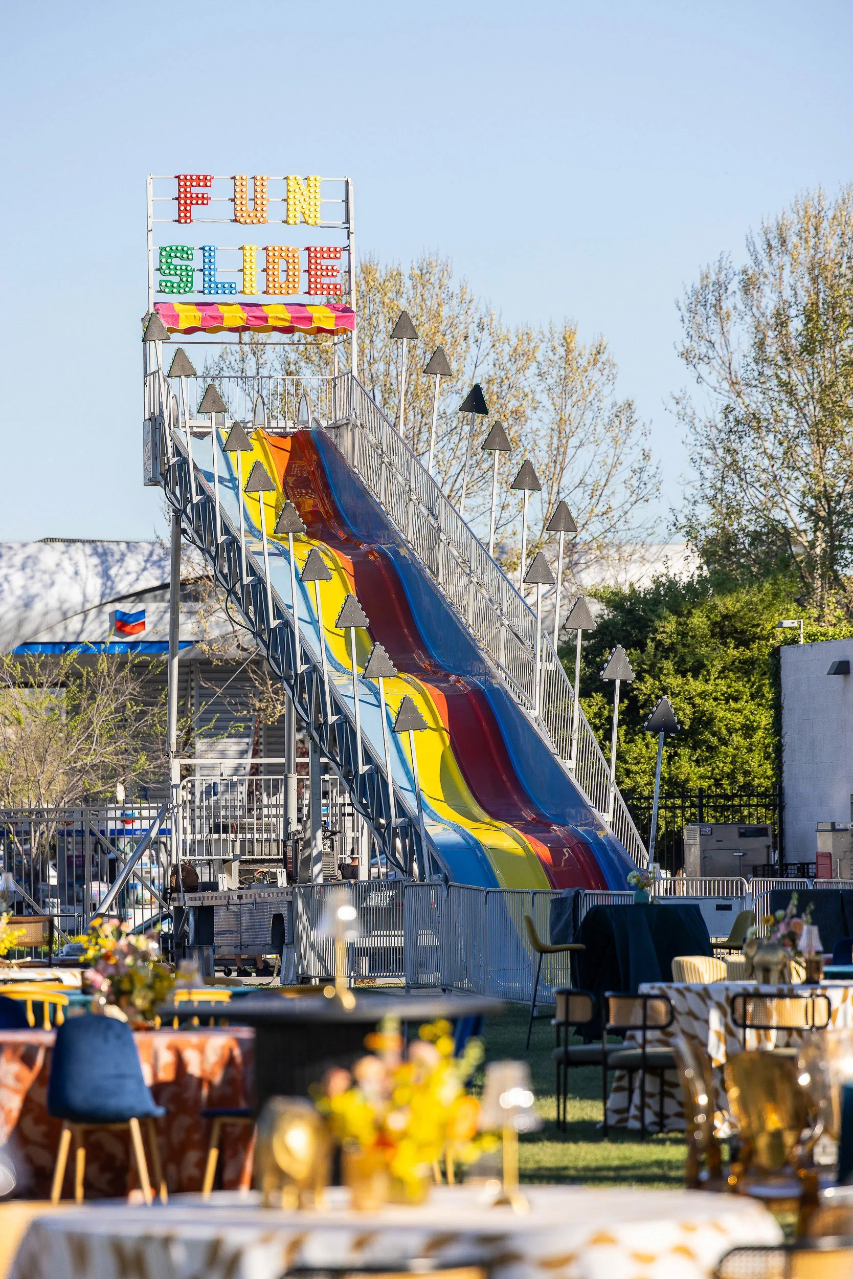 A colorful amusement park slide with a sign that reads 'FUN SLIDE' in large, colorful letters, set against a clear blue sky with trees in the background. There are tables and chairs with decorations in the foreground.