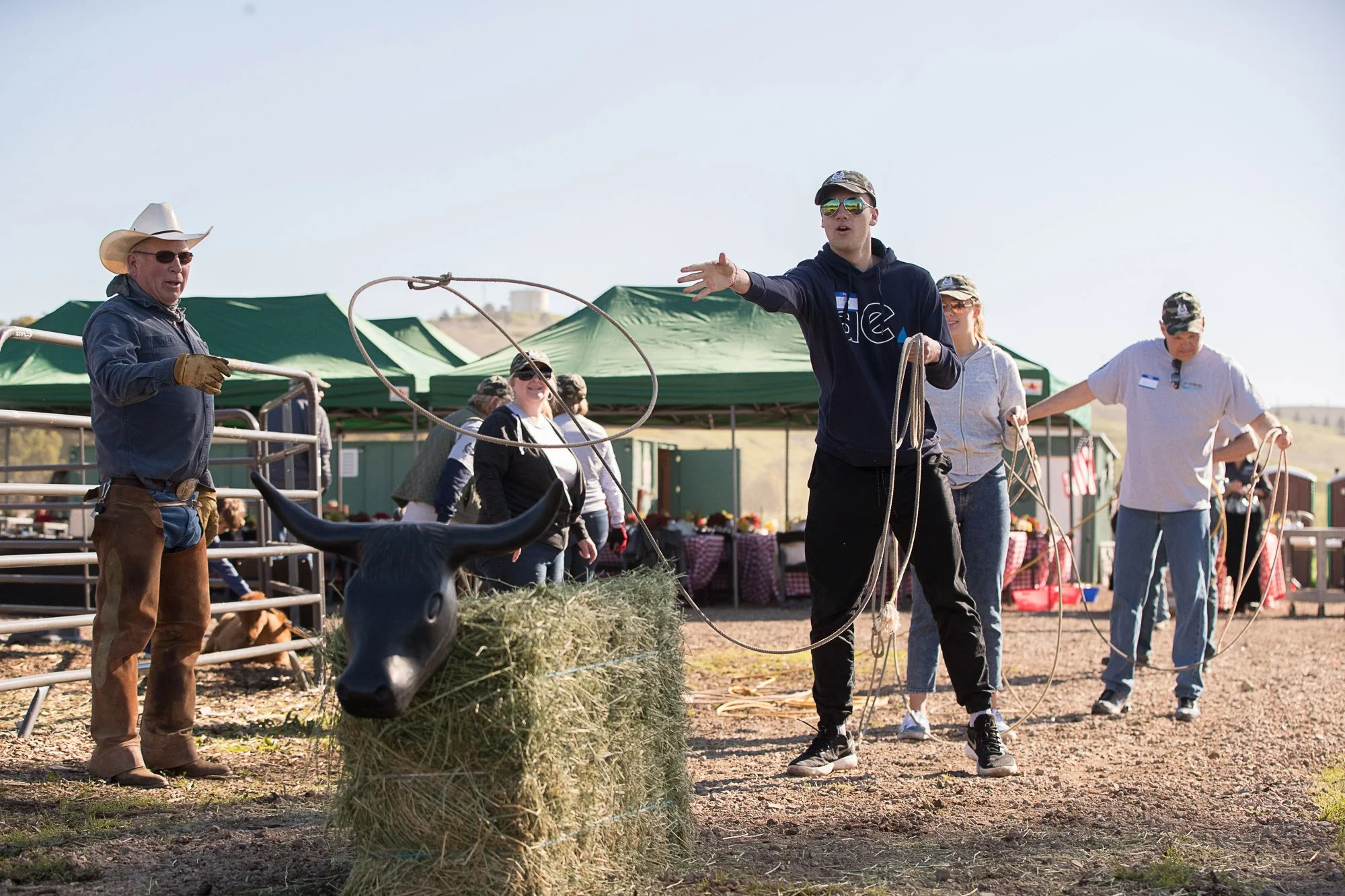 People participating in a ranch or rodeo event outdoors, including a man in a cowboy hat and sunglasses, and a young man holding a rope, with tents and tables set up in the background.