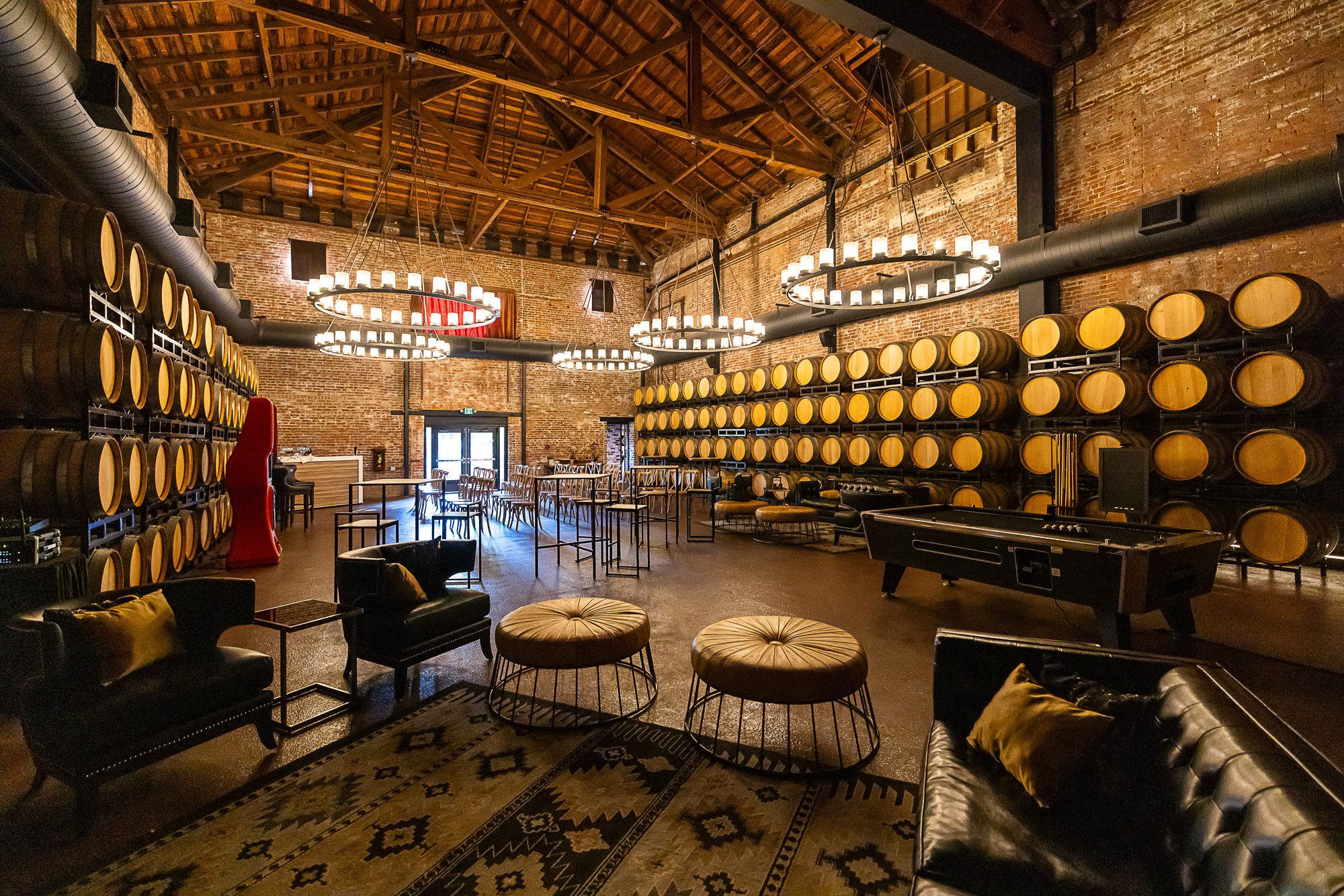 Interior of a rustic venue with brick walls and a high wooden ceiling, decorated with circular chandeliers made of candles, and rows of wine barrels on both sides. Lounge seating and tables are arranged in the space.