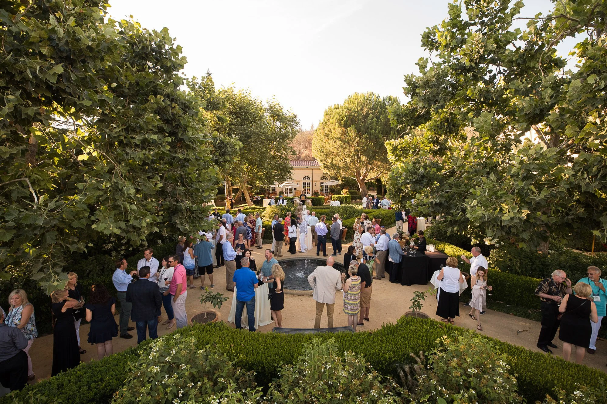 Gathering of people at an outdoor event in a garden with lush trees and shrubbery, a fountain, and a large house in the background, with the scenic view framed by two large leafy trees.