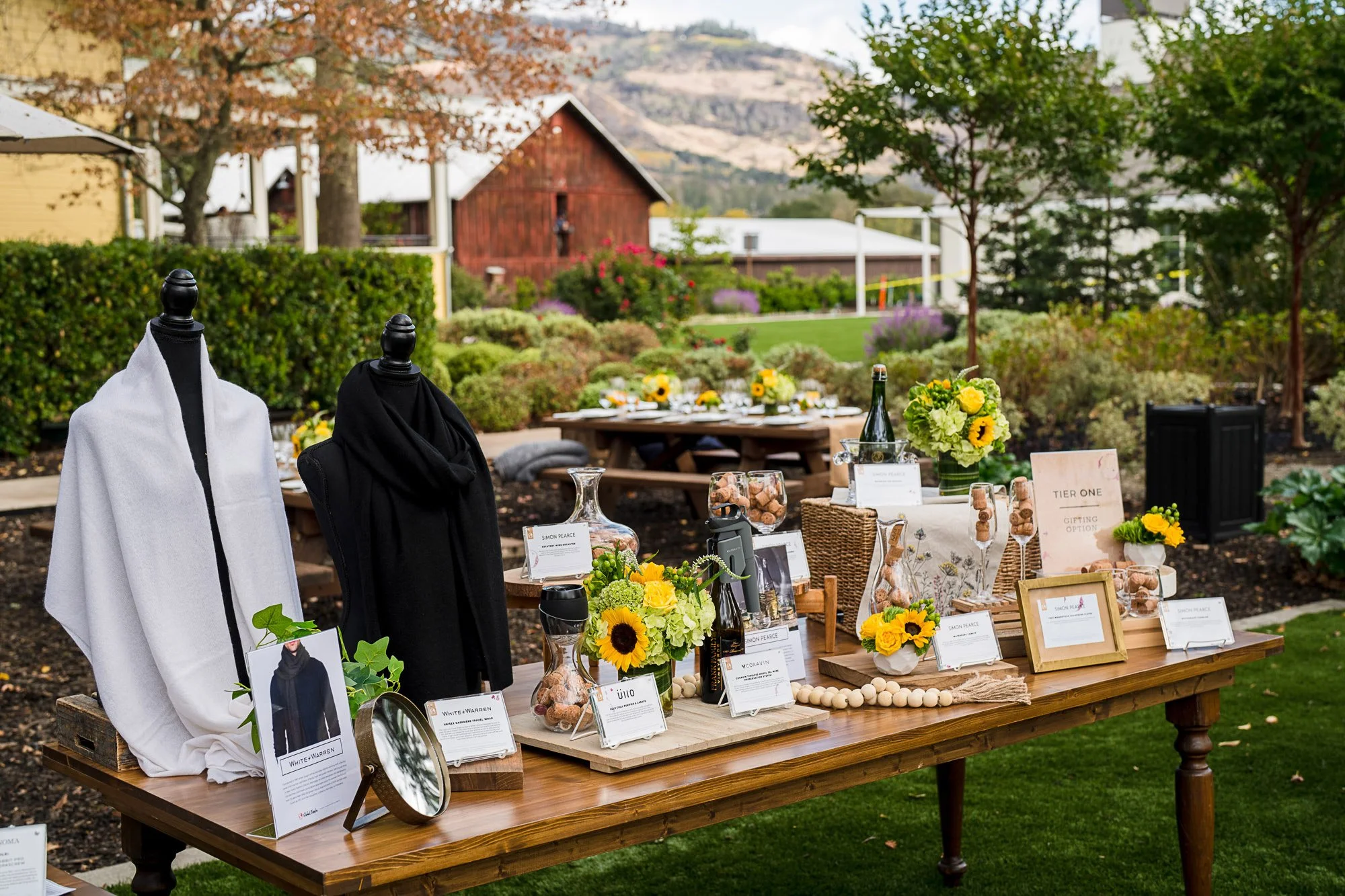 An outdoor event setup with tables displaying various products, including wine bottles, framed pictures, and signs with descriptions. The tables are decorated with sunflower and green floral arrangements. Mannequins are draped with black and white cl