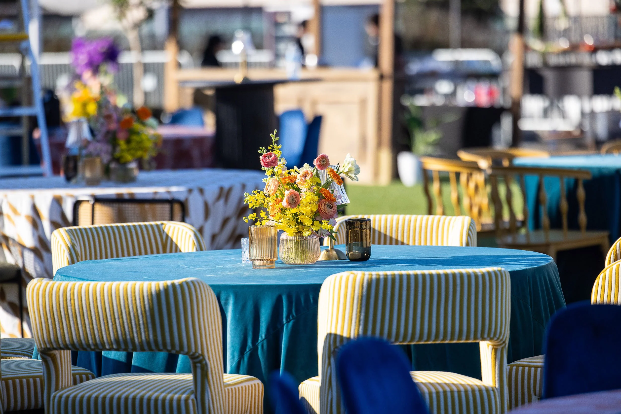 Outdoor dining area with a table covered in a turquoise tablecloth, adorned with a colorful flower arrangement in a glass vase. Surrounding chairs have yellow and white striped cushions. Background includes blurred view of additional tables, chairs, 