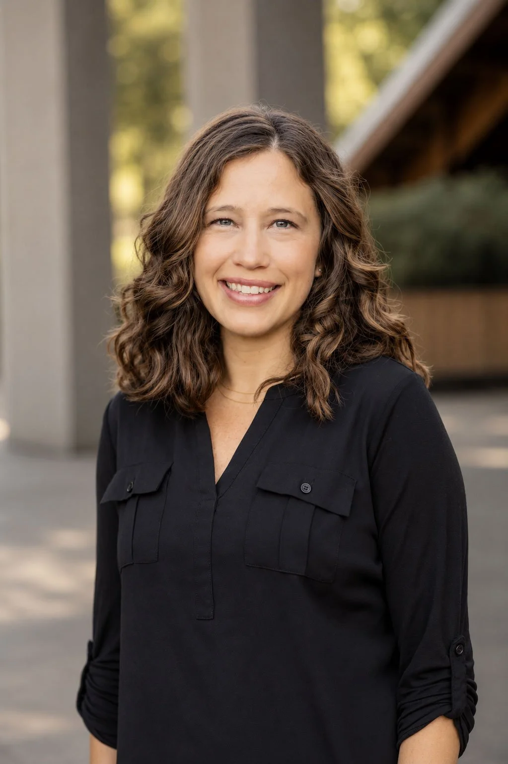 A woman with shoulder-length curly brown hair, wearing a black button-up shirt, smiling outdoors with blurred trees and structures in the background.