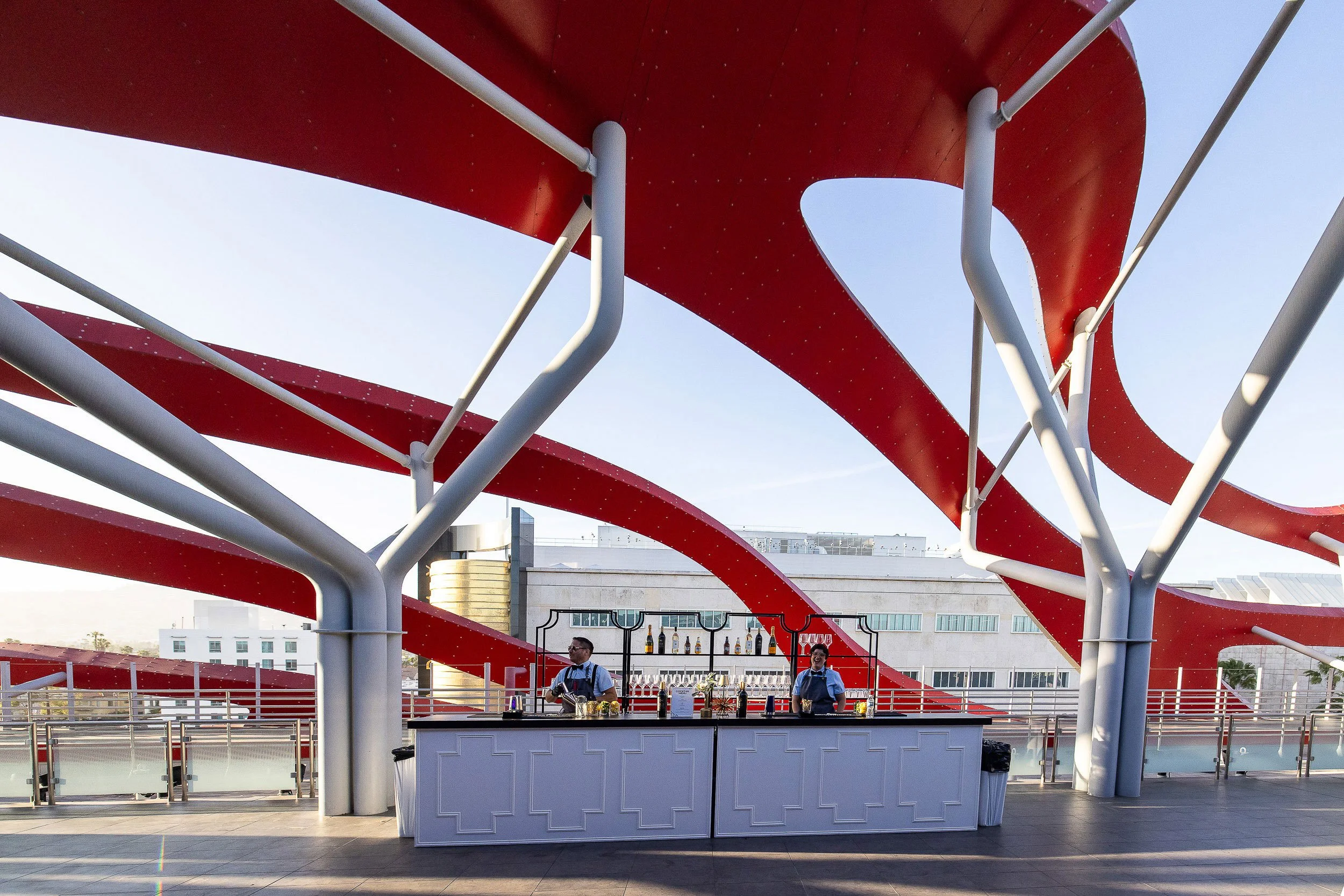 Outdoor bar with two bartenders under a large red architectural canopy with curved shapes and white structural supports.