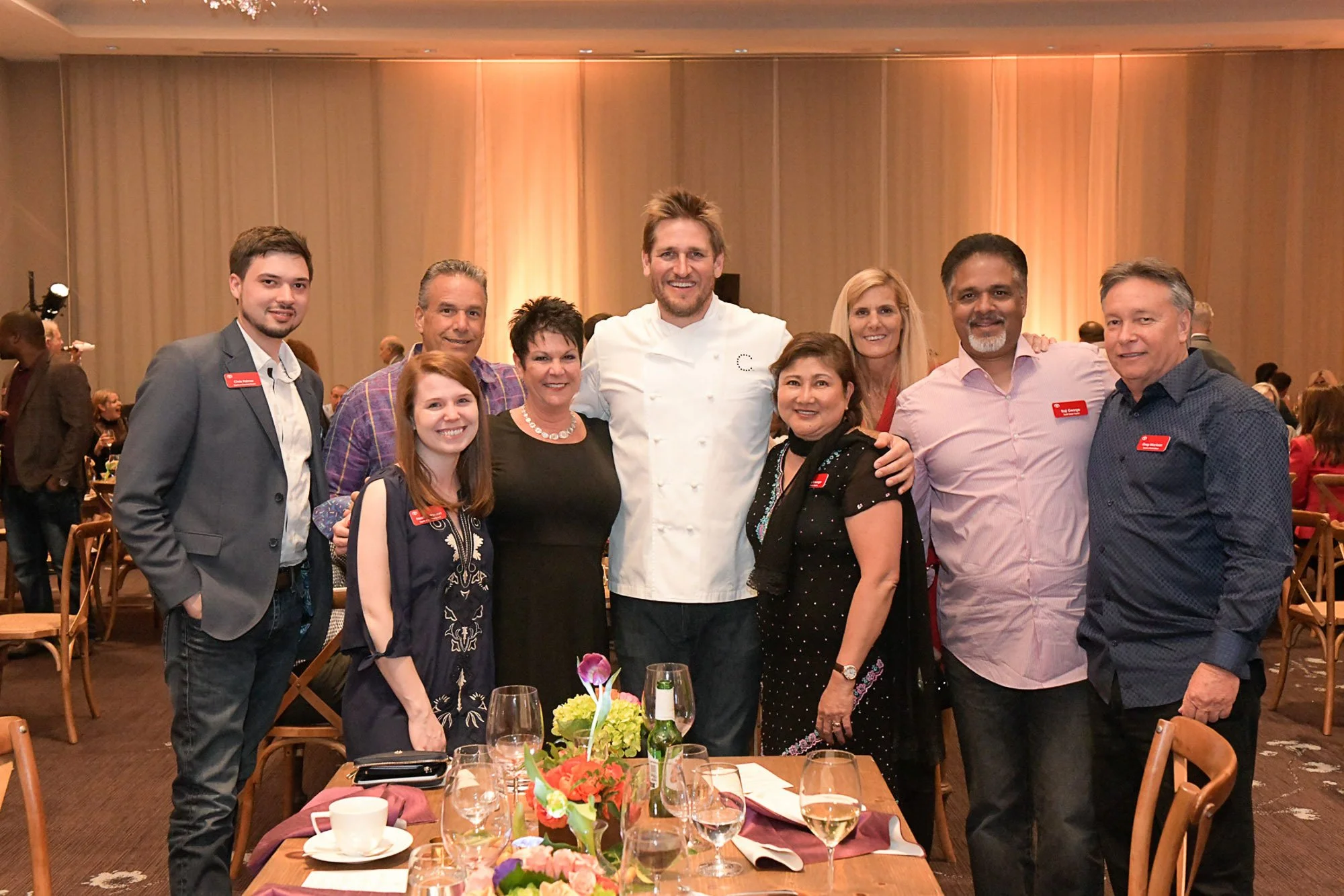 Group of nine people, including a chef in white, posing together at a banquet table with floral centerpiece, glasses, and tableware in a banquet hall with warm lighting and beige curtains.